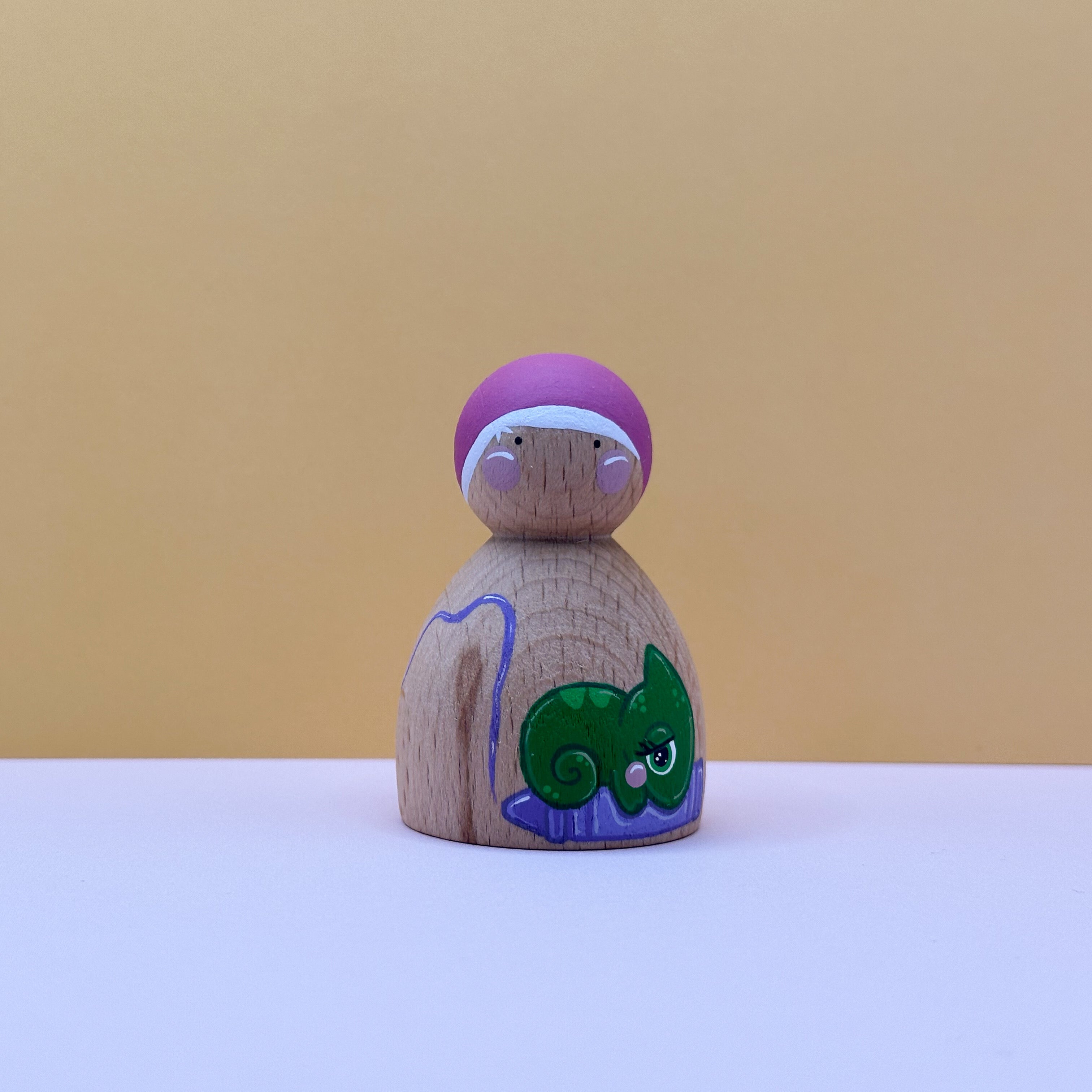Wooden peg doll with a purple hat and green lizard design on a beige background