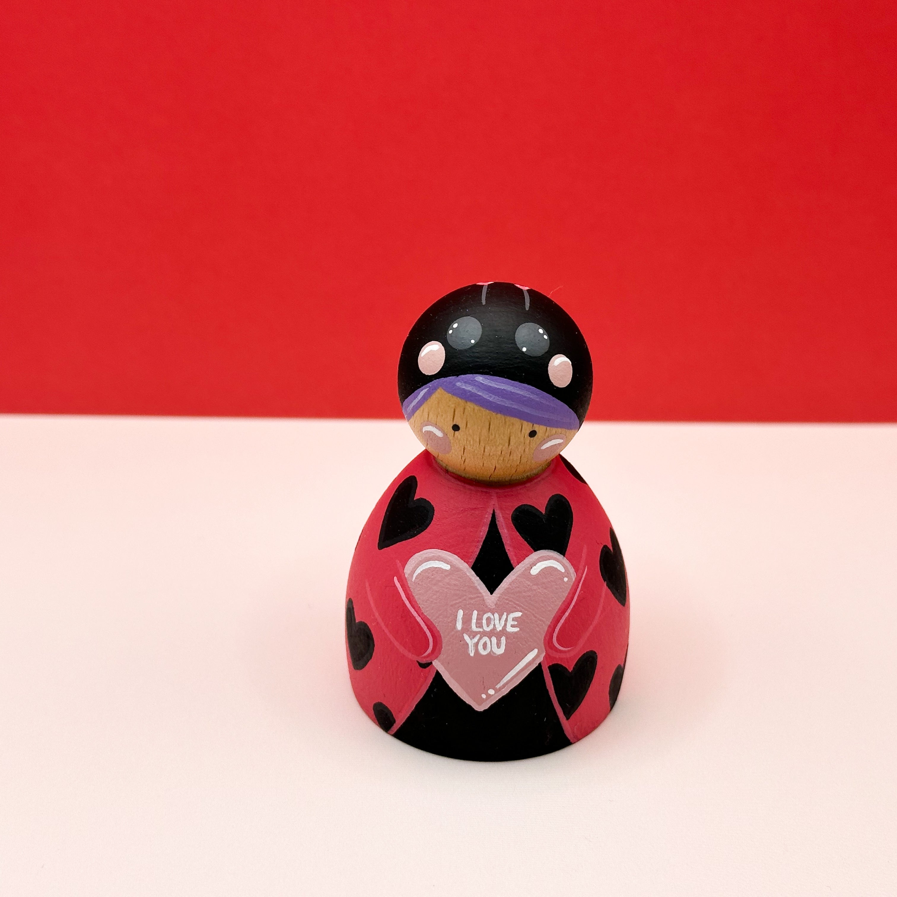 a chubby wooden peg doll dressed as a ladybird holding a love heart on a red background