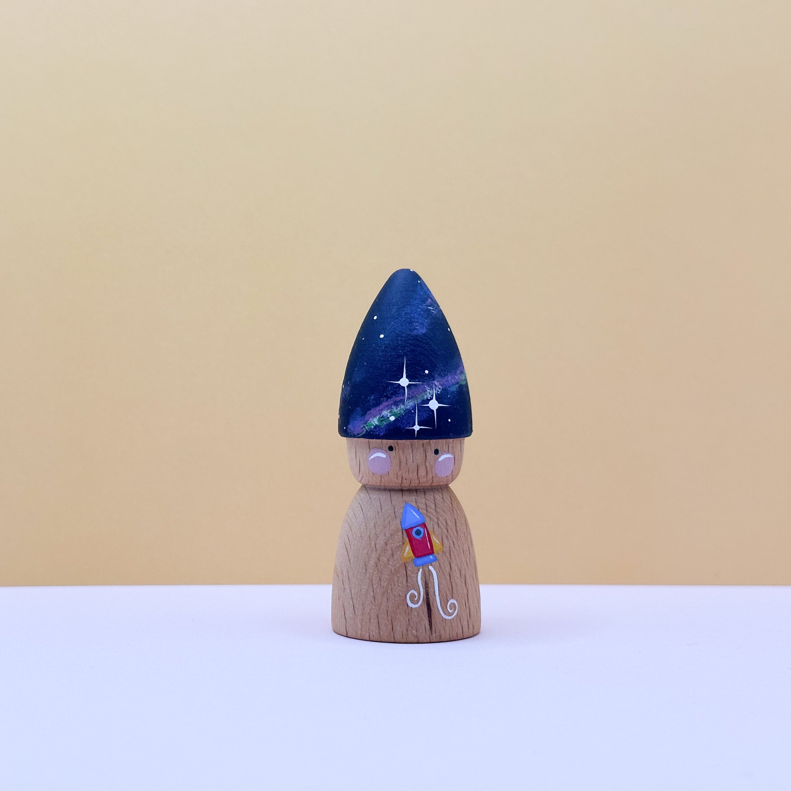 Wooden peg doll with a blue hood and rocket ship on a beige background