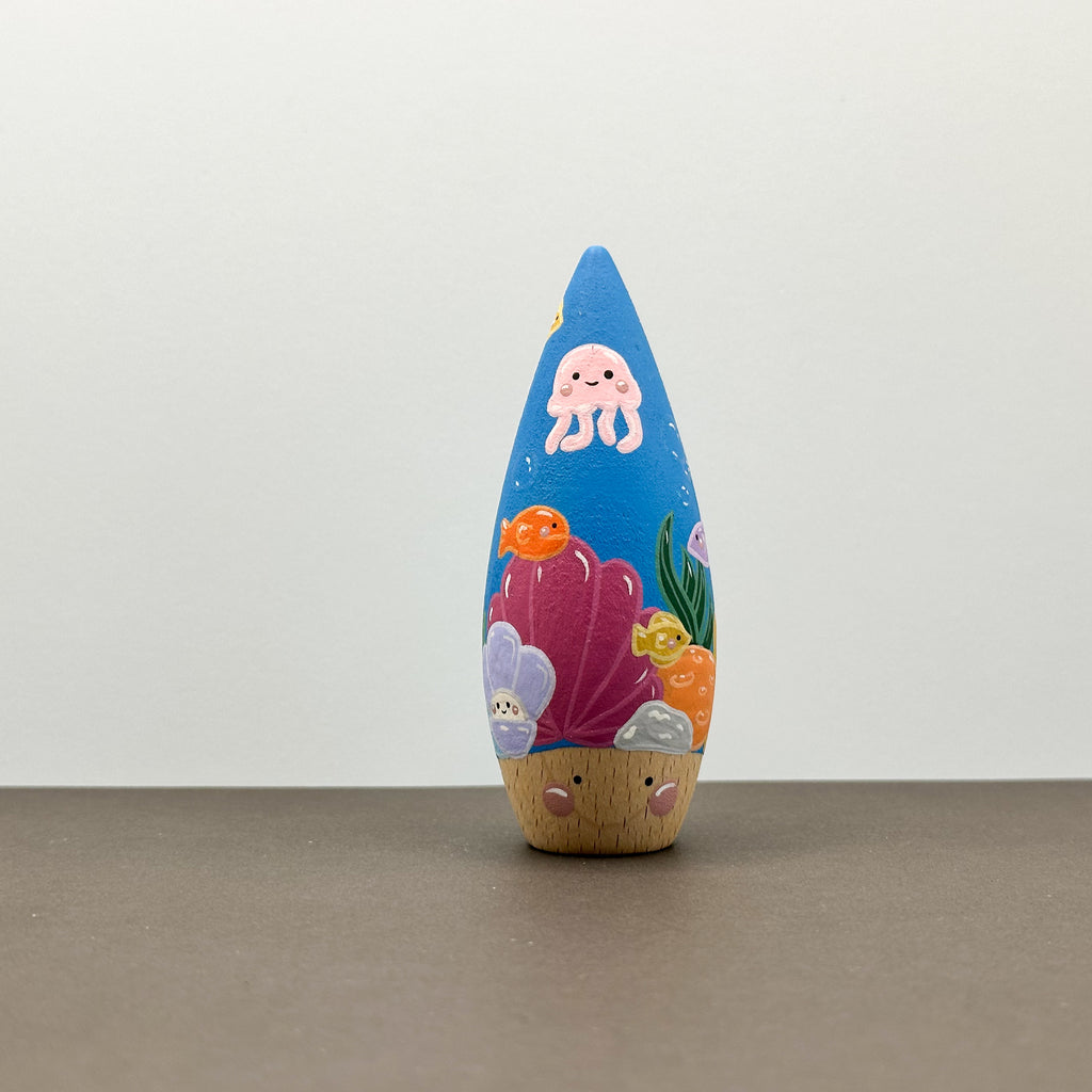 A wooden tear drop shaped toy with a coral reef theme on a grey background. 