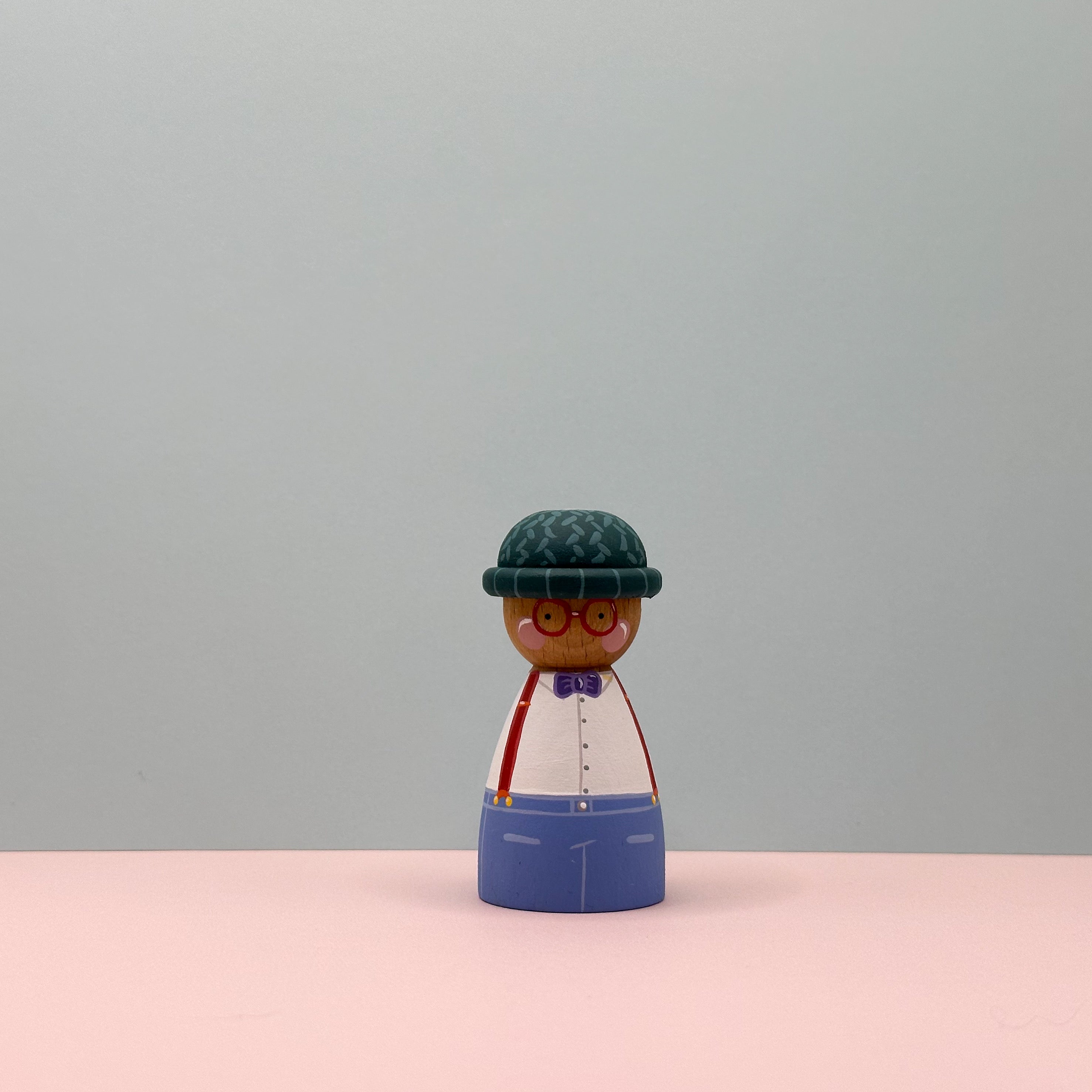 Wooden peg doll with a green hat, white shirt, blue pants, and red braces on a pink surface with gray background