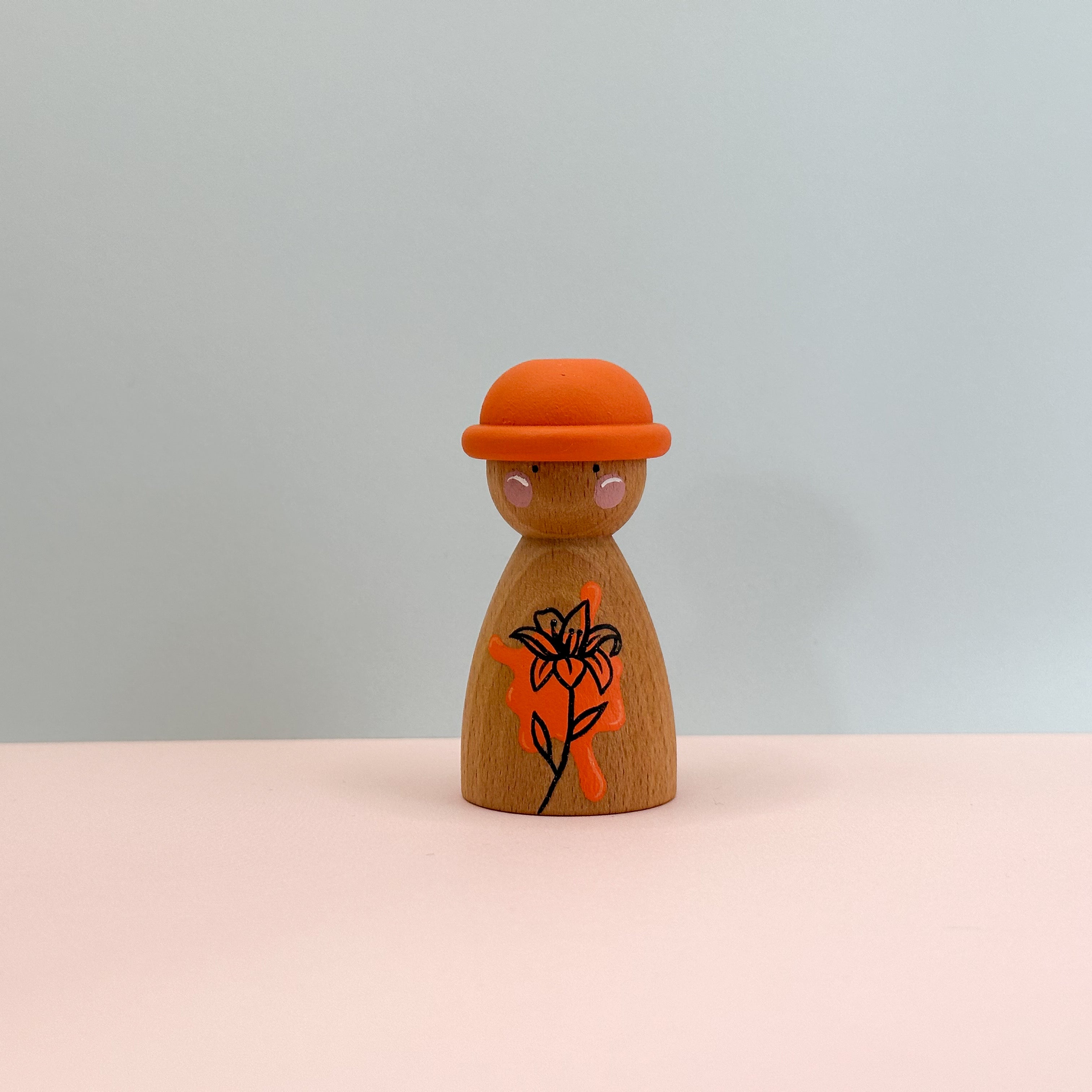 Wooden peg doll with an orange hat and black floral design on a light gray background