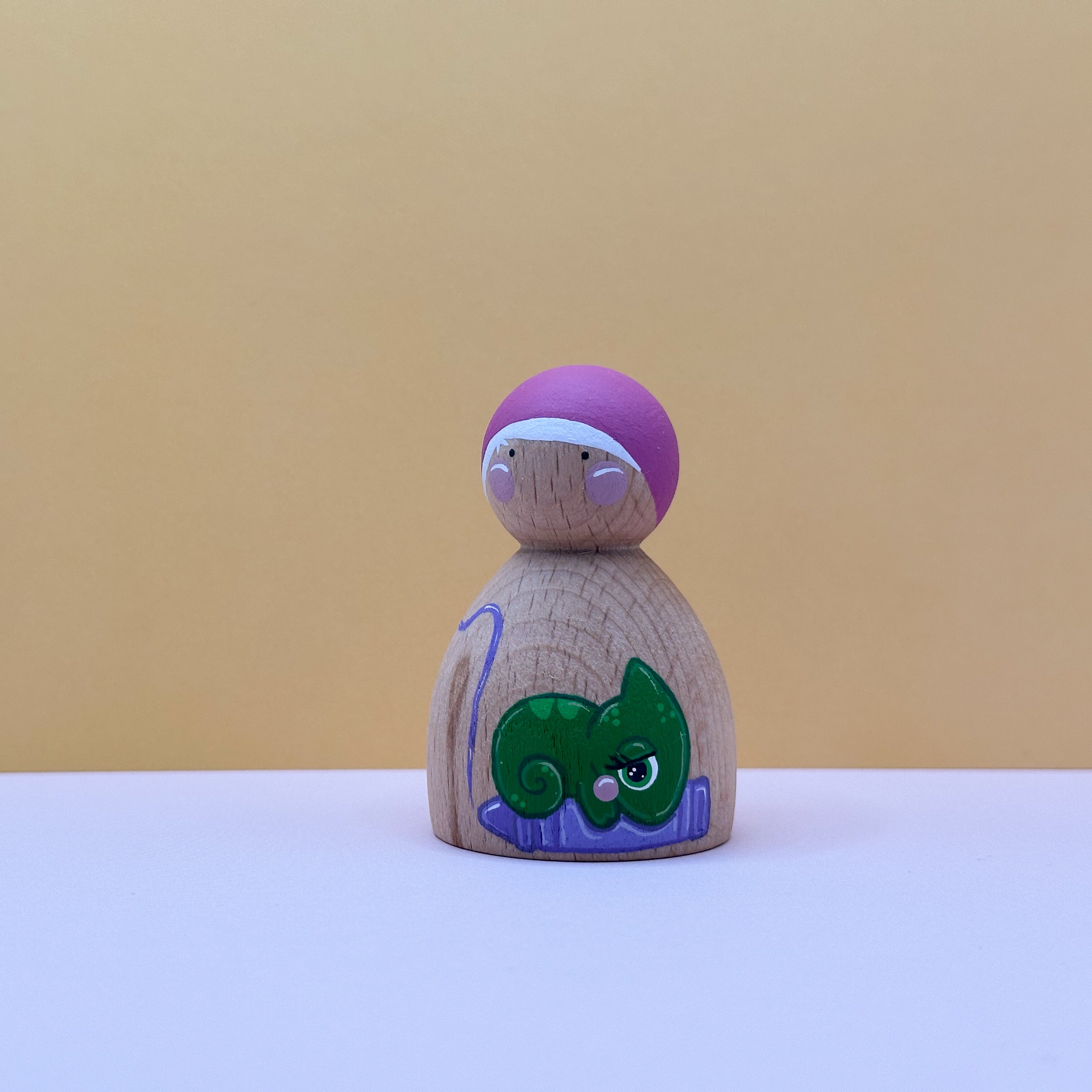Wooden peg doll with a purple top and green lizard design on a beige background