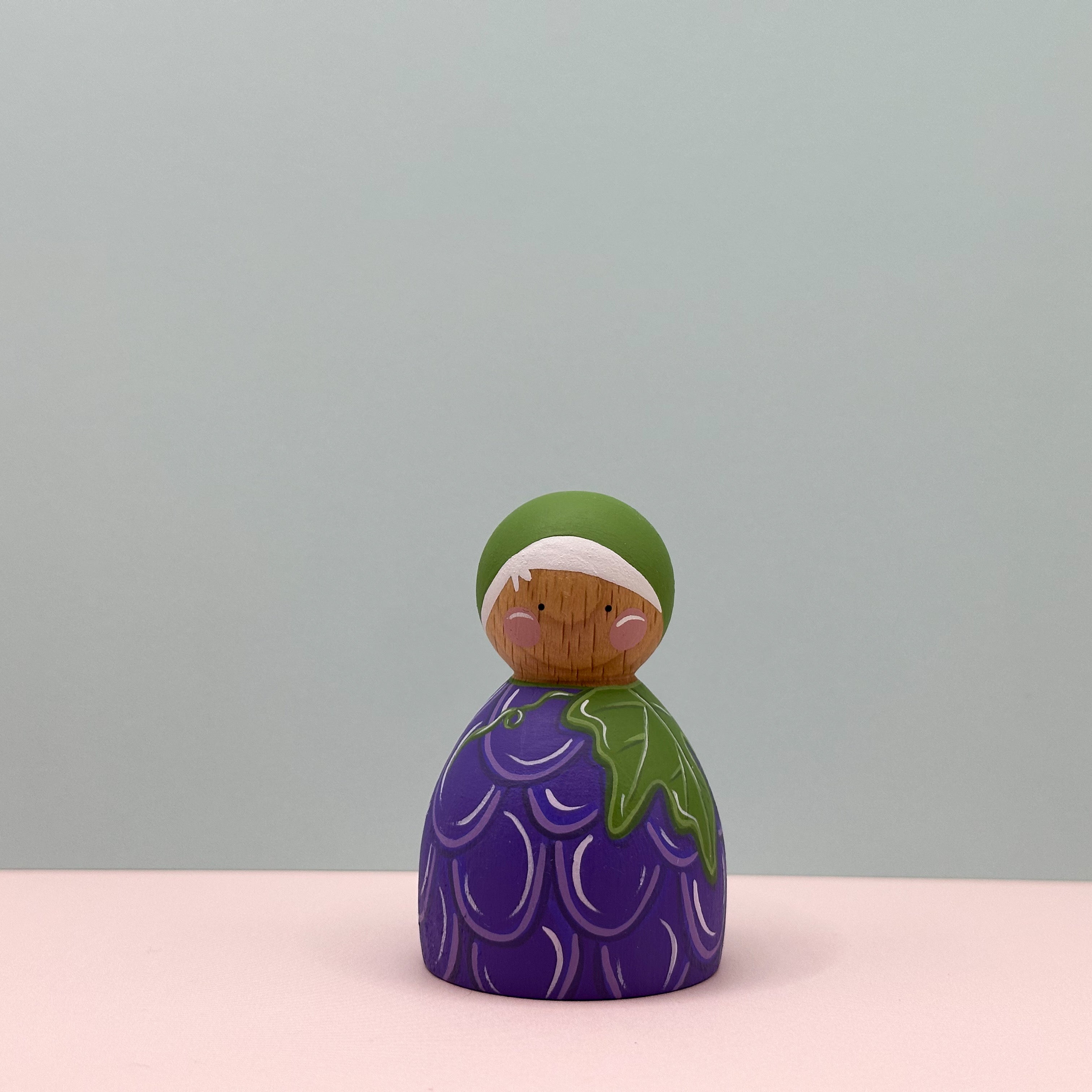 Decorative figurine with purple grape body and green top on a light pink surface and gray background