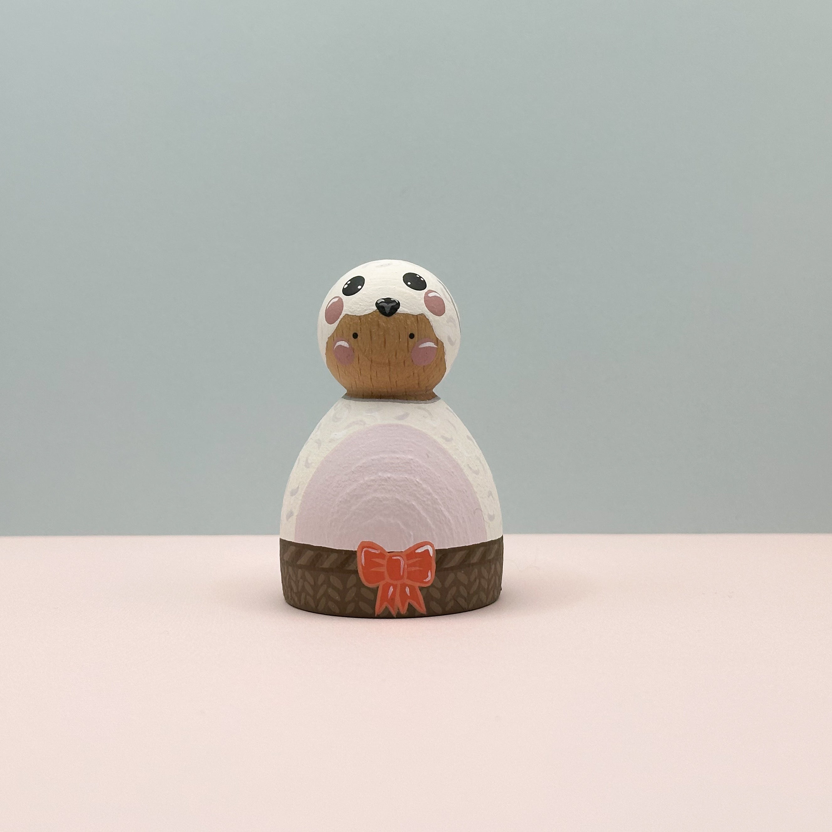 Small wooden figurine with a white face, brown body, and pink bow on a light gray background