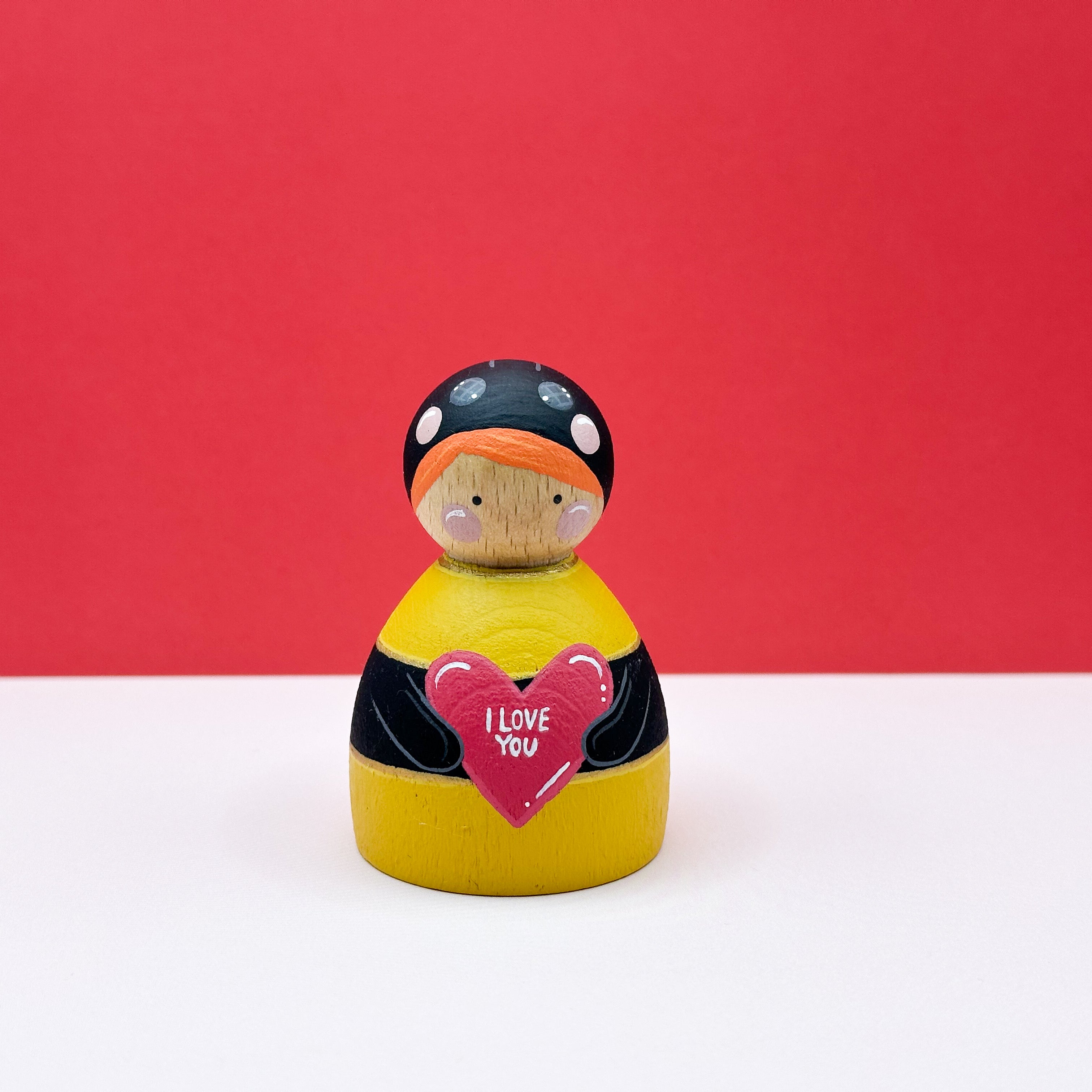 a chubby wooden peg doll dressed as a bee holding a love heart on a red background