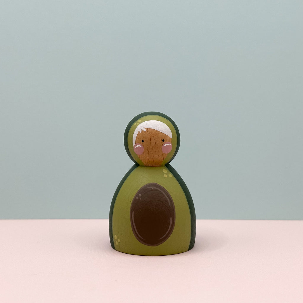 Wooden toy avocado with a face on a light pink surface and light blue background