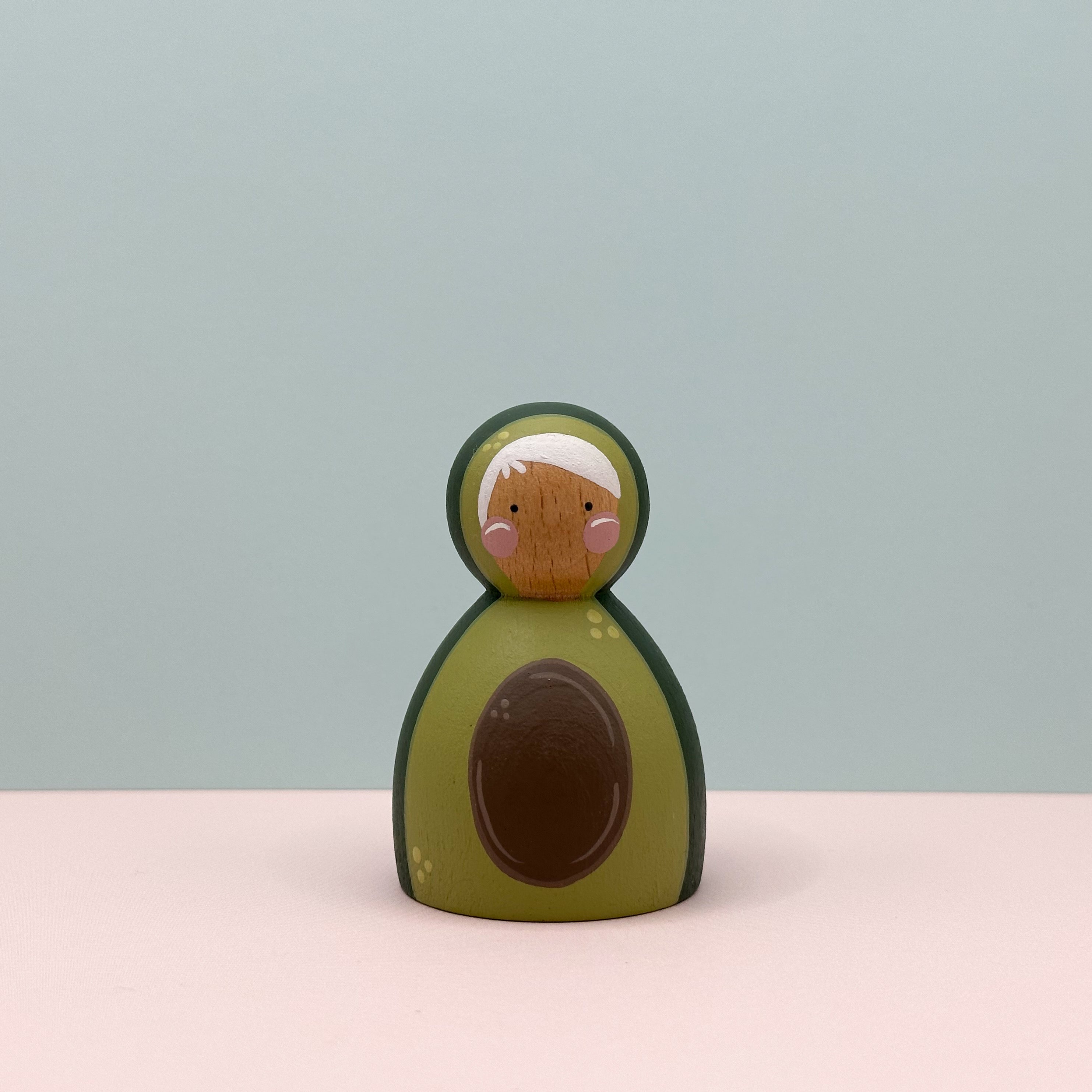 Wooden toy avocado with a face on a light pink surface and light blue background