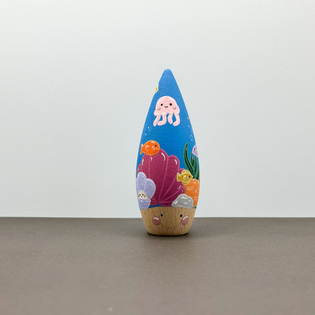 A wooden tear drop shaped toy with a coral reef theme on a grey background. 