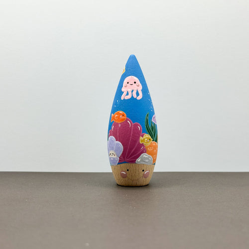 A wooden tear drop shaped toy with a coral reef theme on a grey background. 