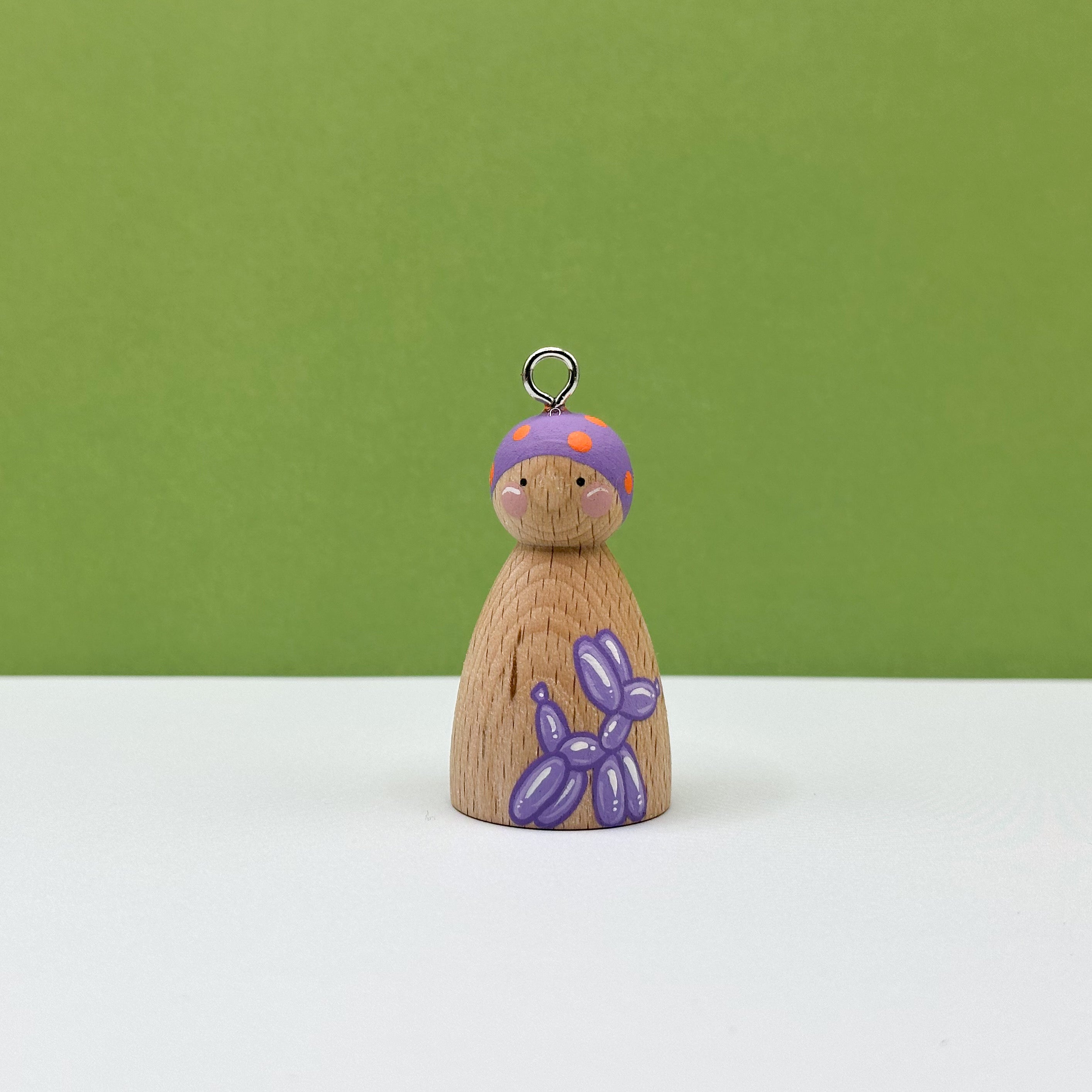 Wooden peg doll with a purple hat and balloon dog accessory on a green background
