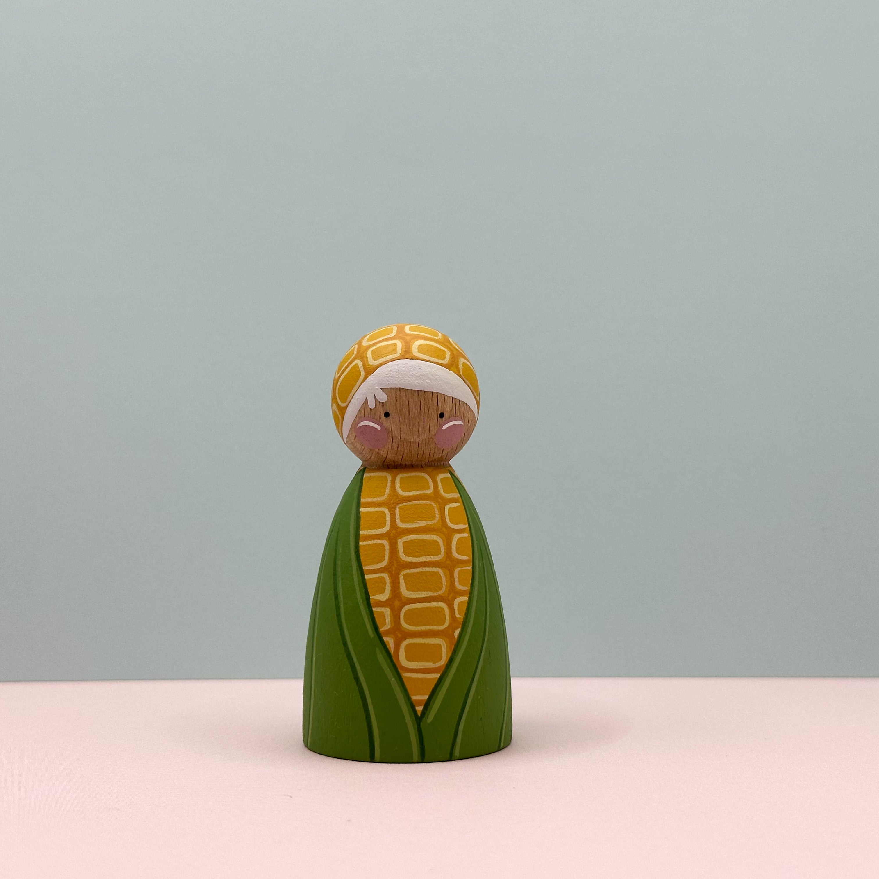Wooden figurine of a corn character with a patterned headband on a light pink surface and light gray background