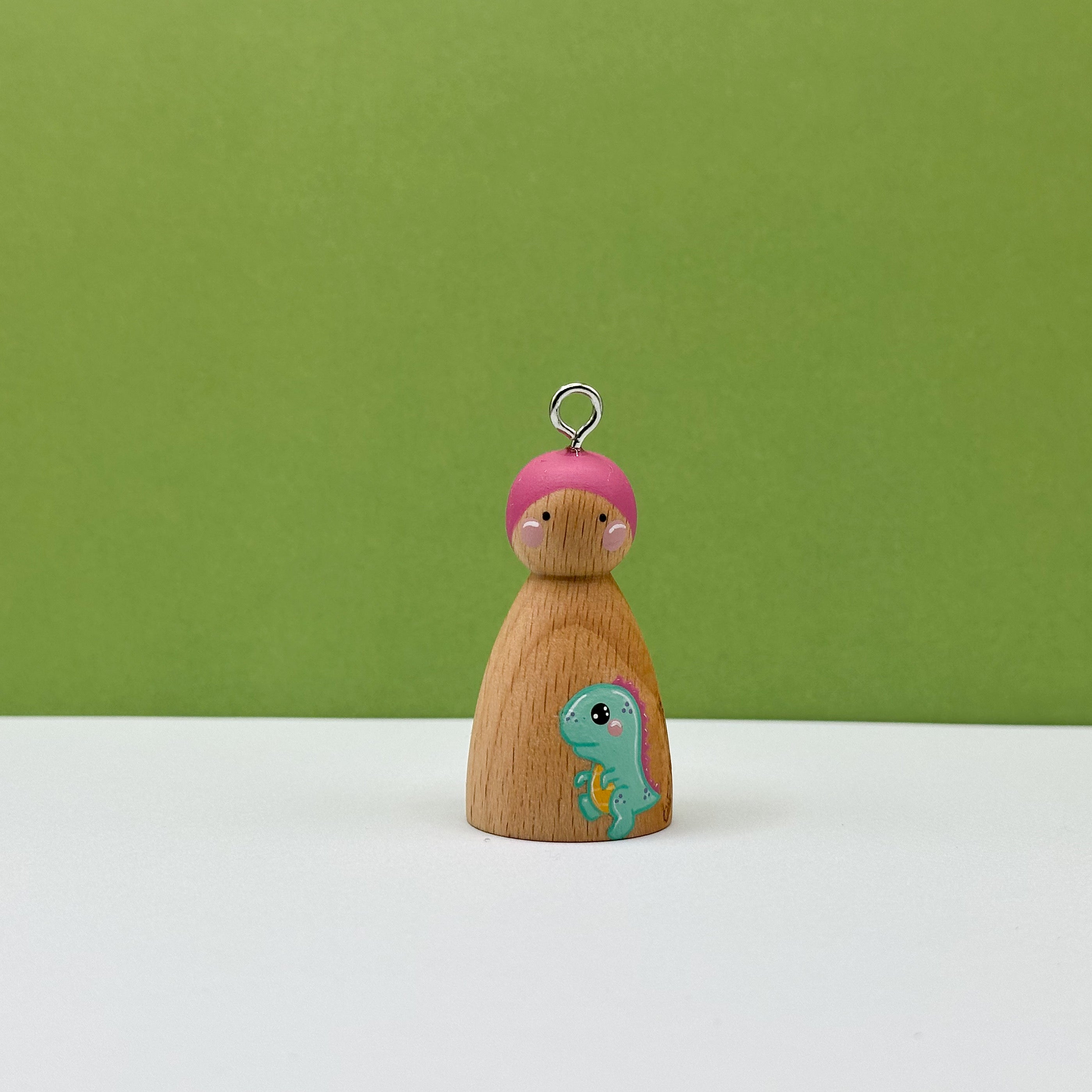 Wooden peg doll with pink hair and a dinosaur design on a green background