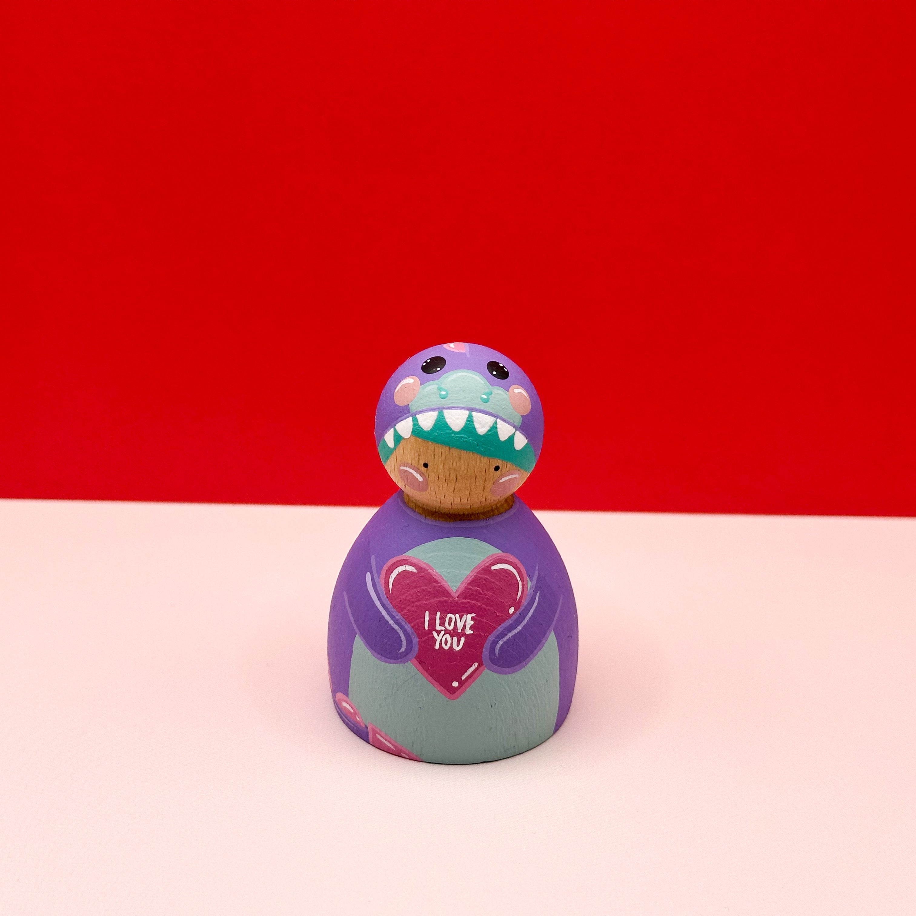 a chubby wooden peg doll dressed as a purple dinosaur holding a love heart on a red background