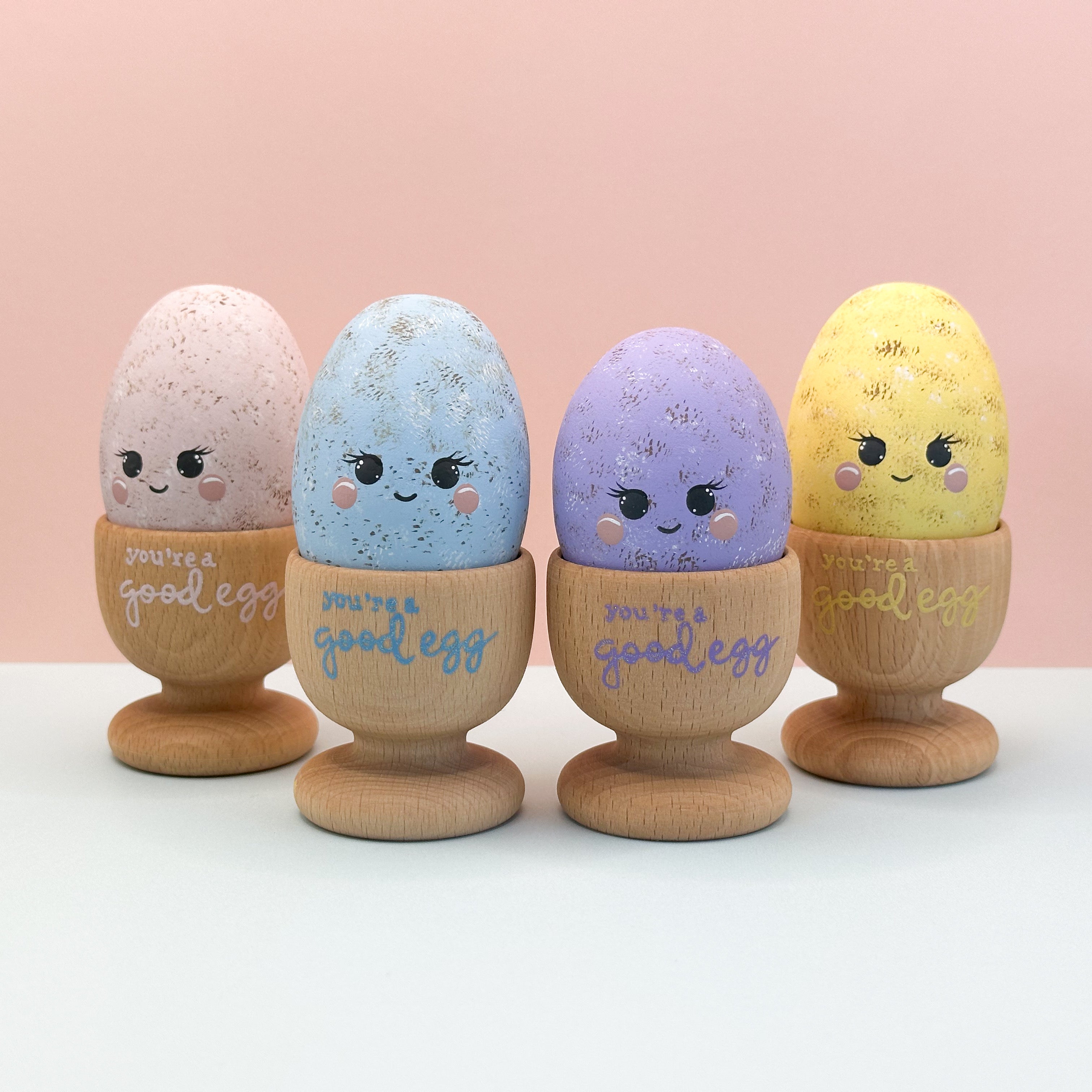 Four decorative eggs with faces and 'you're a good egg' text on wooden stands against a pink background.