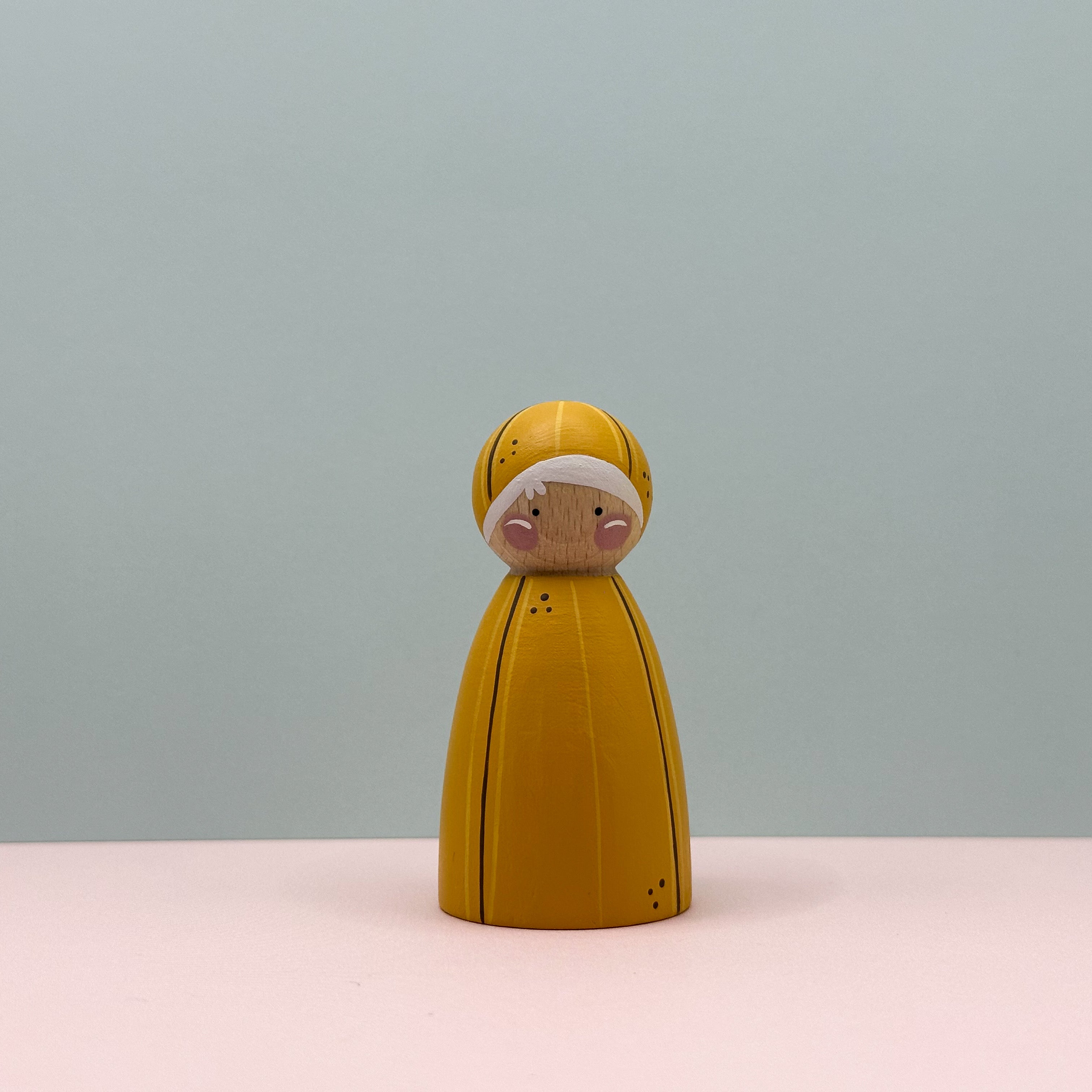 Wooden peg doll with a yellow banana dress on a light pink surface and light gray background