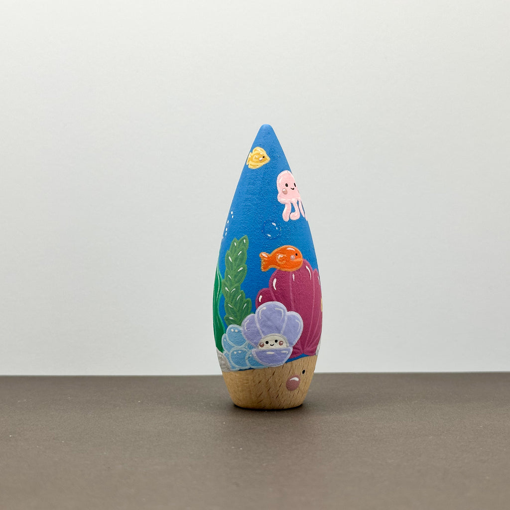 A wooden tear drop shaped toy with a coral reef theme on a grey background. 