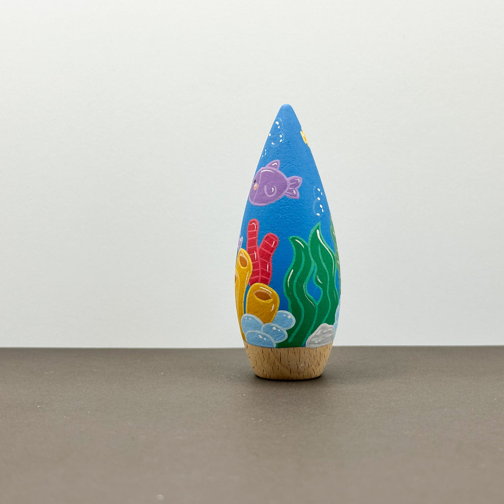 A wooden tear drop shaped toy with a coral reef theme on a grey background. 