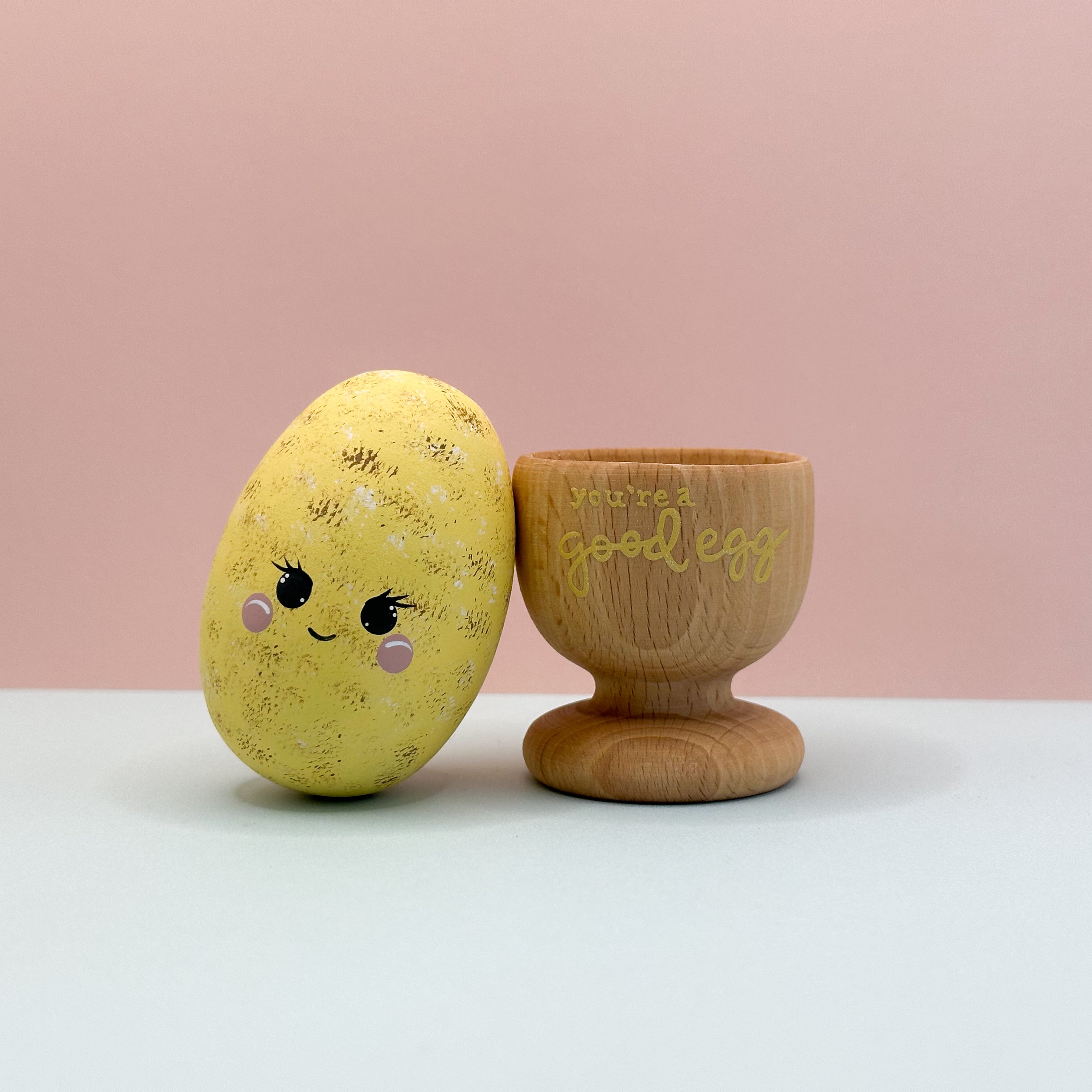 Yellow decorative egg with a face and wooden egg cup on a pink background