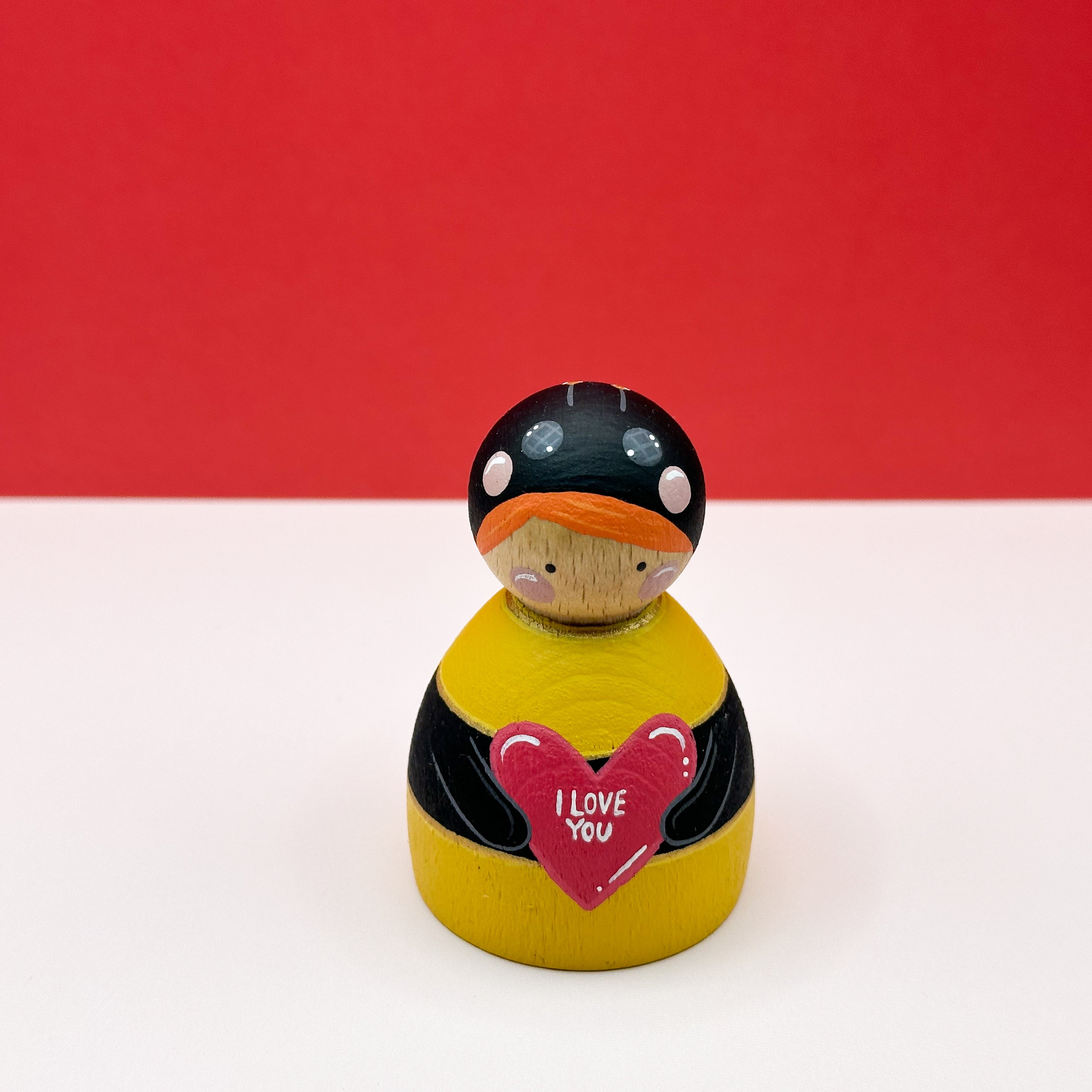 a chubby wooden peg doll dressed as a bee holding a love heart on a red background