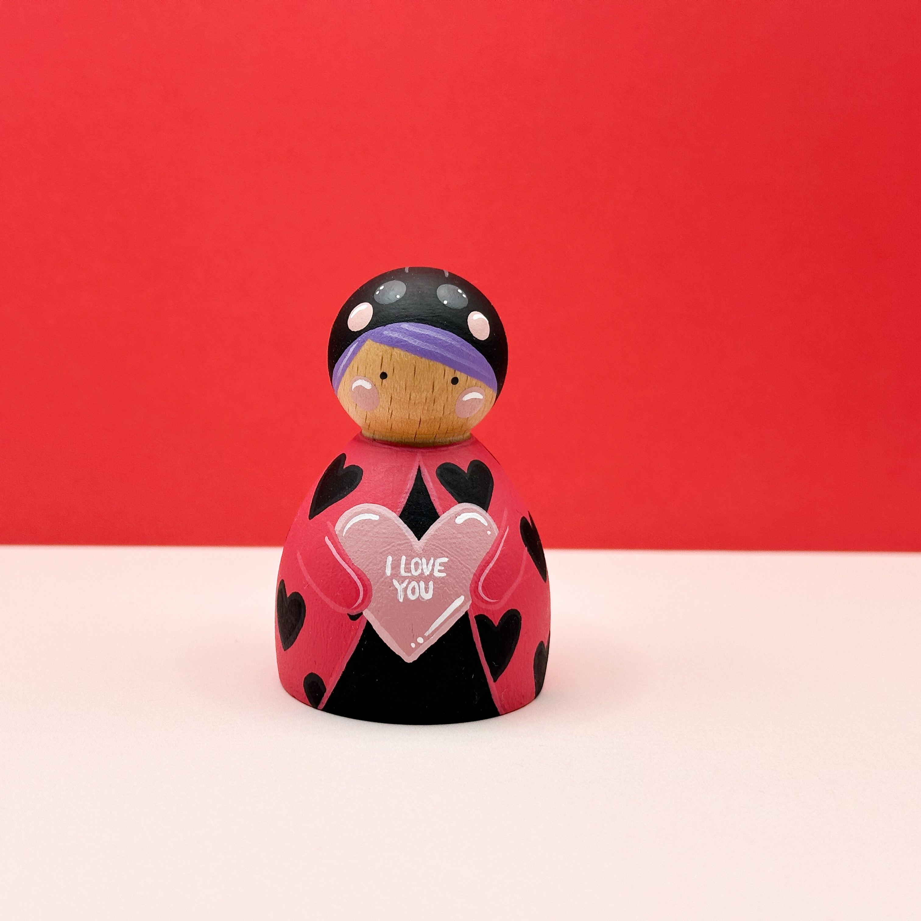 a chubby wooden peg doll dressed as a ladybird holding a love heart on a red background