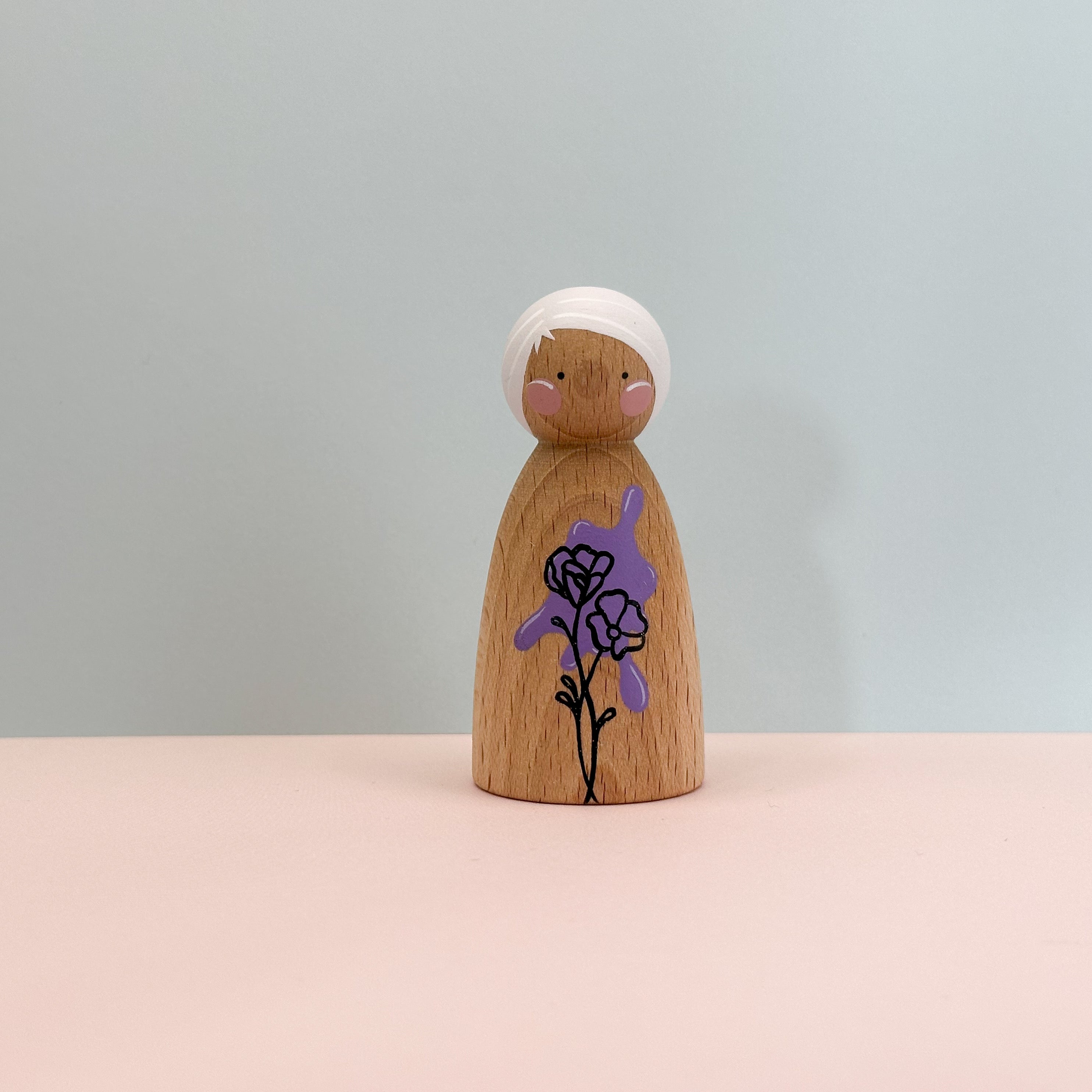 Wooden peg doll with a white headband and purple flower design on a light gray background