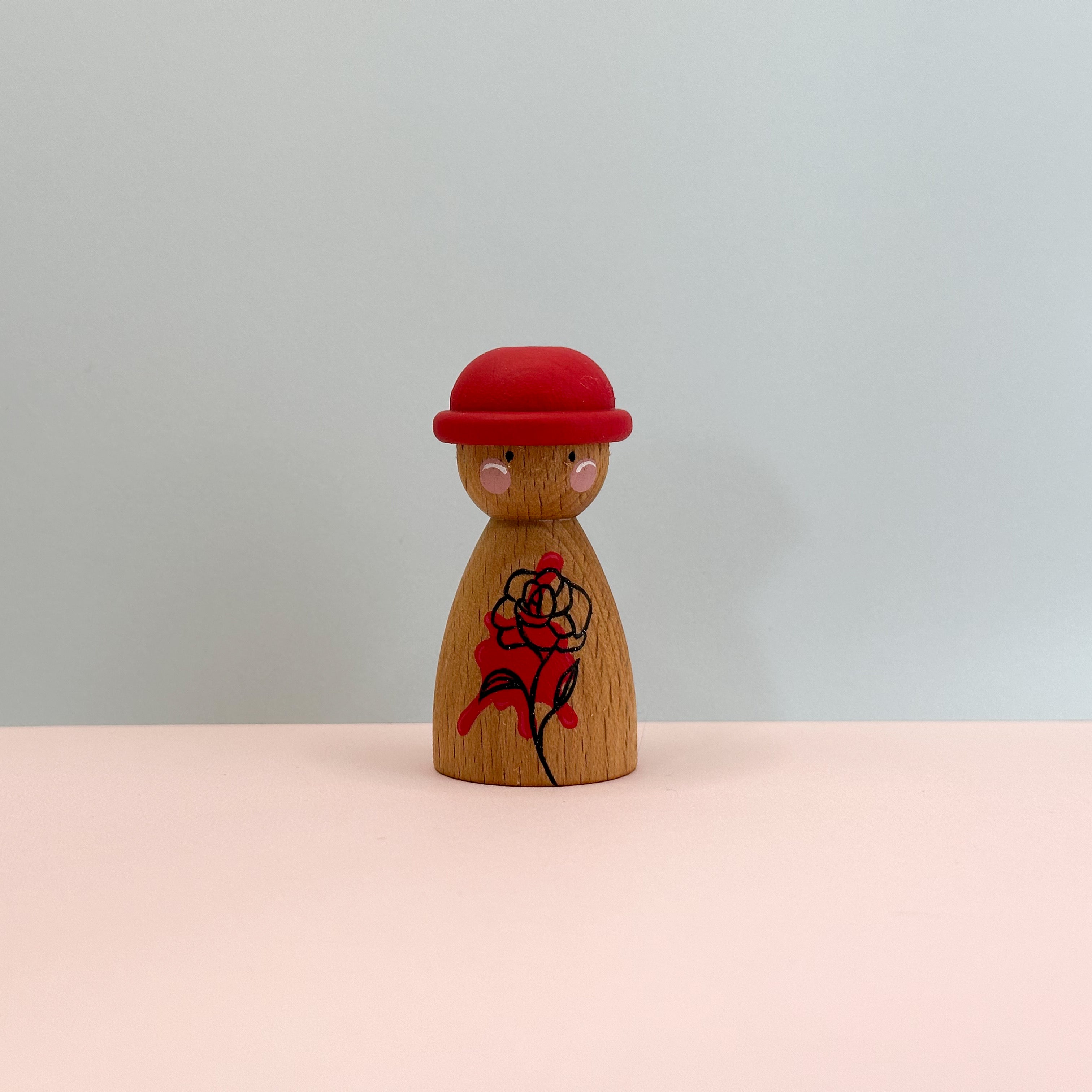 Wooden peg doll with a red hat and black and red design on a light gray background