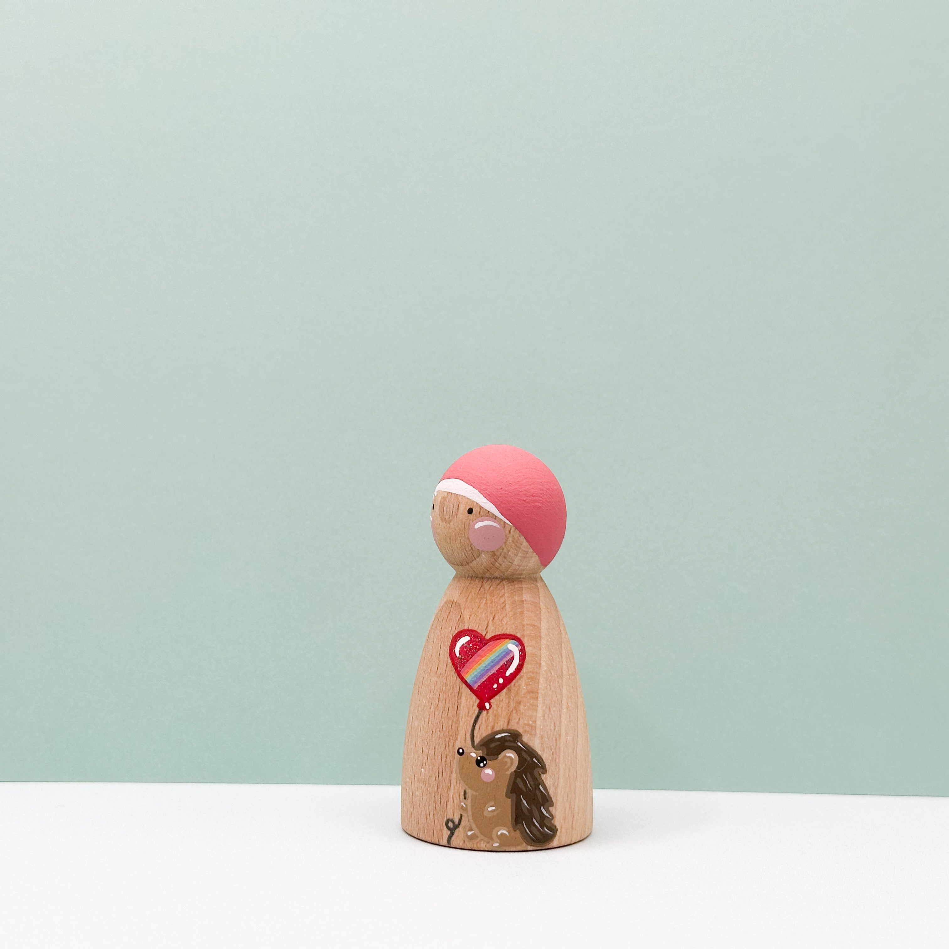 Wooden peg doll with a pink top and heart-shaped balloon on a light green background