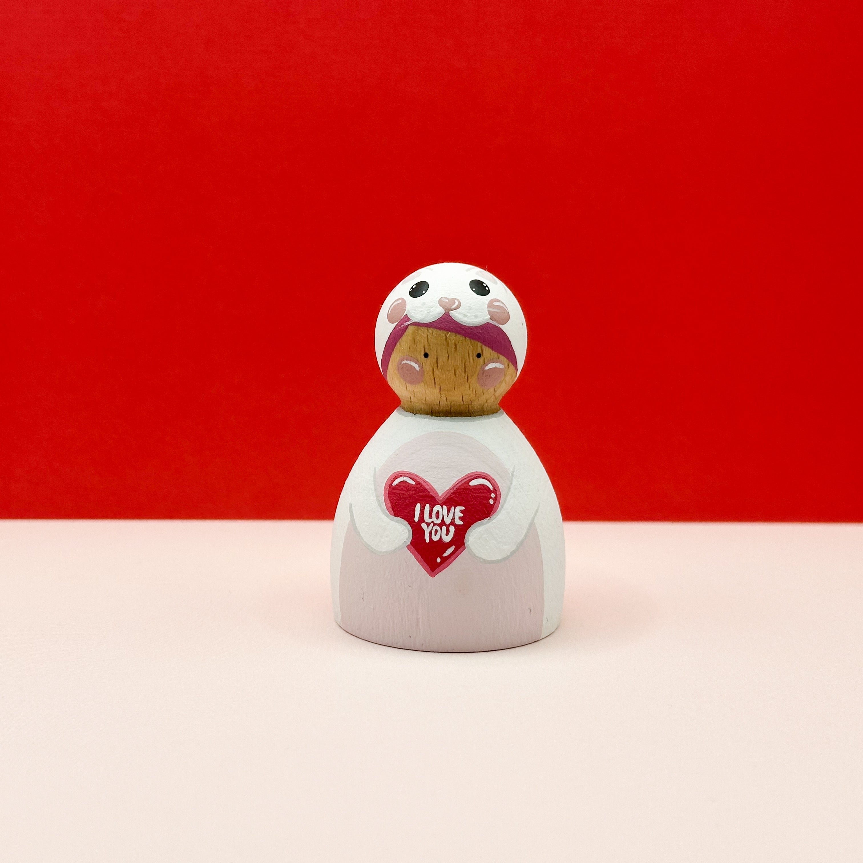 a chubby wooden peg doll dressed as a white cat holding a love heart on a red background