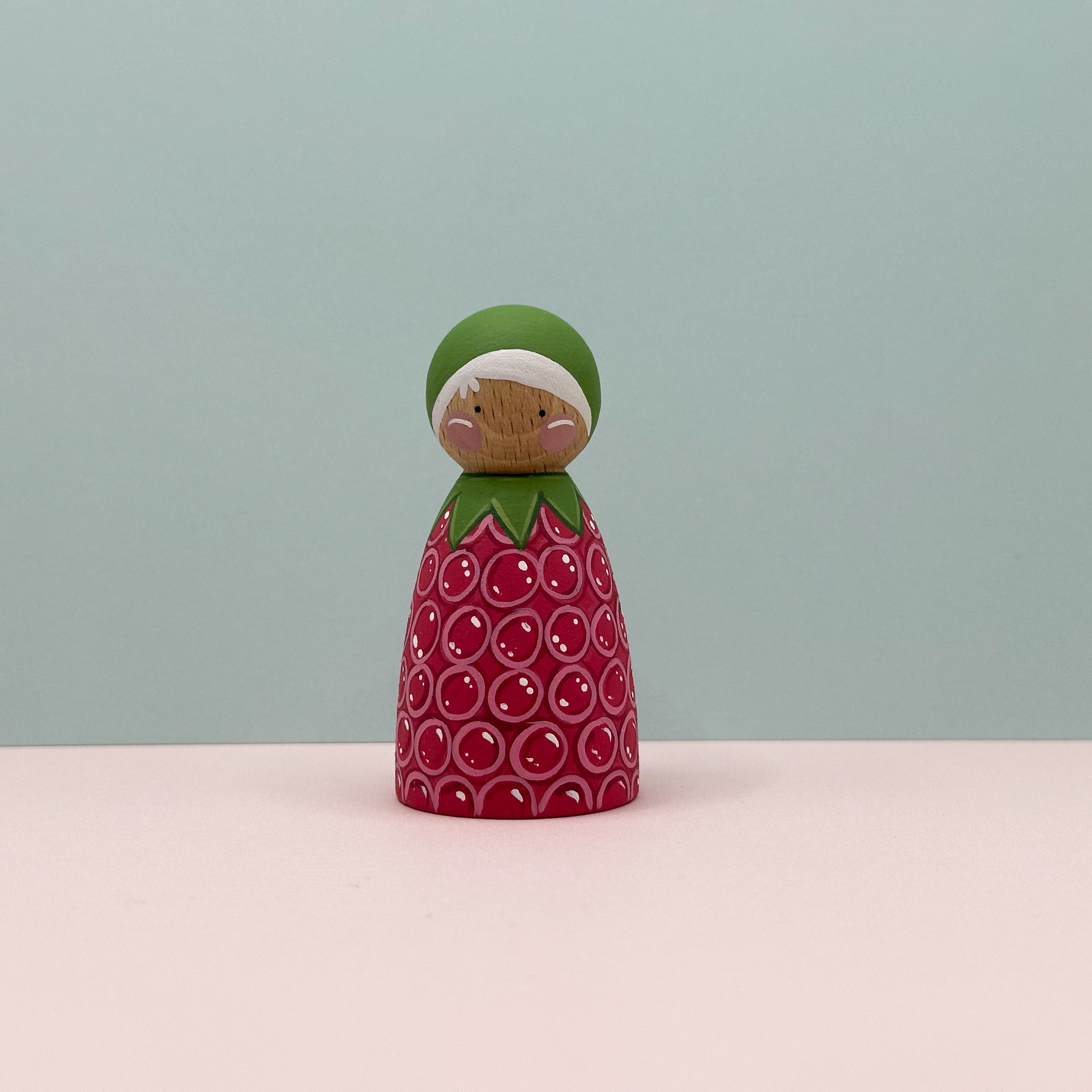 Wooden figurine shaped like a raspberry with a green top on a light pink surface and light blue background
