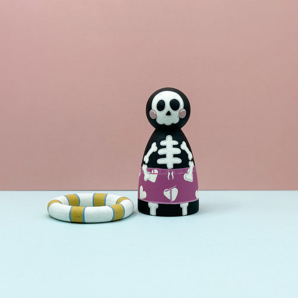 Skeleton figurine with a pink skirt and heart pattern next to a striped ring on a light blue surface with a pink background.