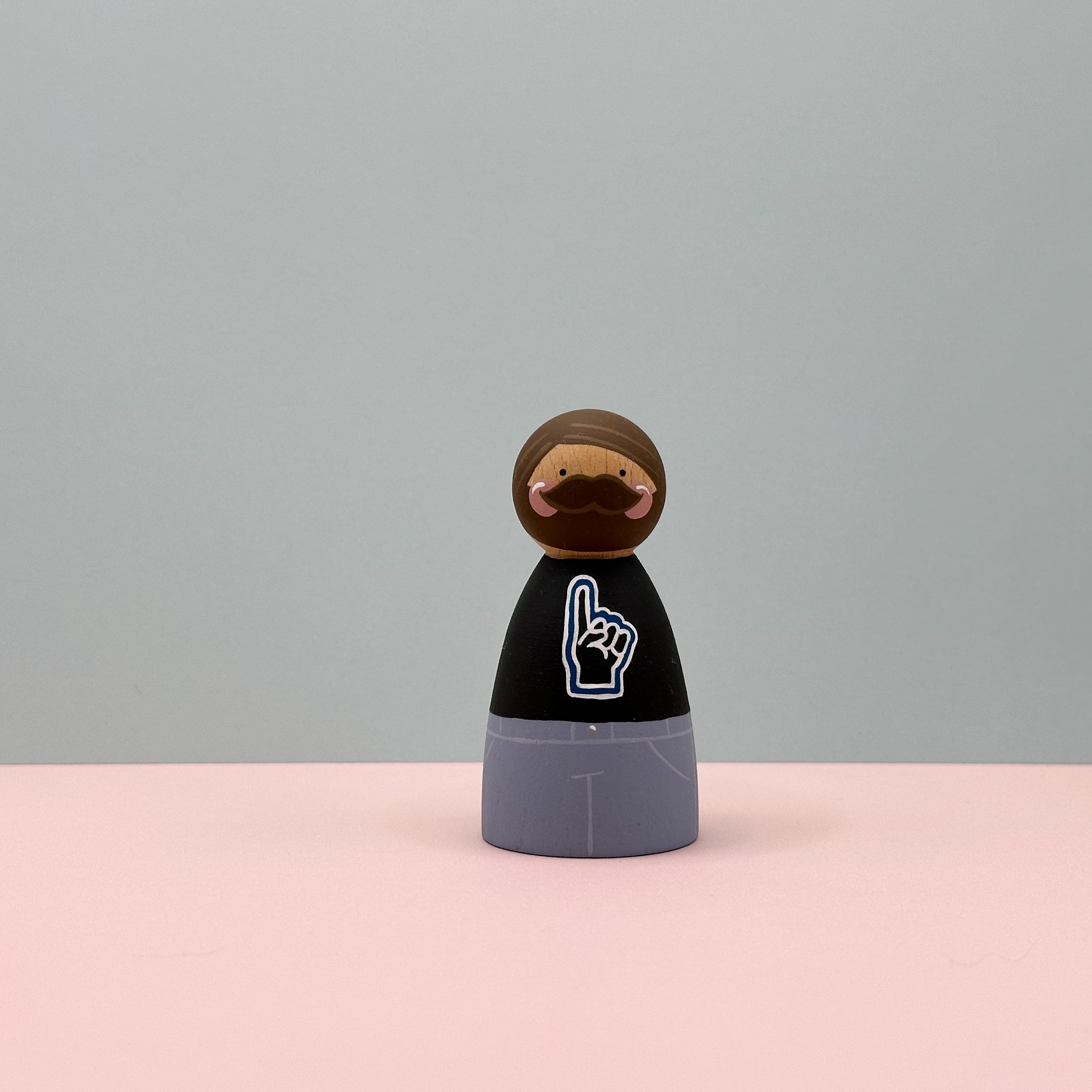 Wooden figurine with a stylized face and hand gesture on a pink and gray background
