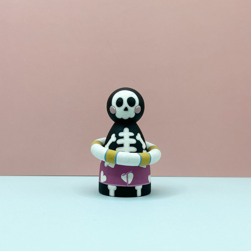 Skeleton figurine with a purple shirt and yellow arm bands on a pink background