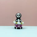 Skeleton figurine with a purple shirt and yellow arm bands on a pink background