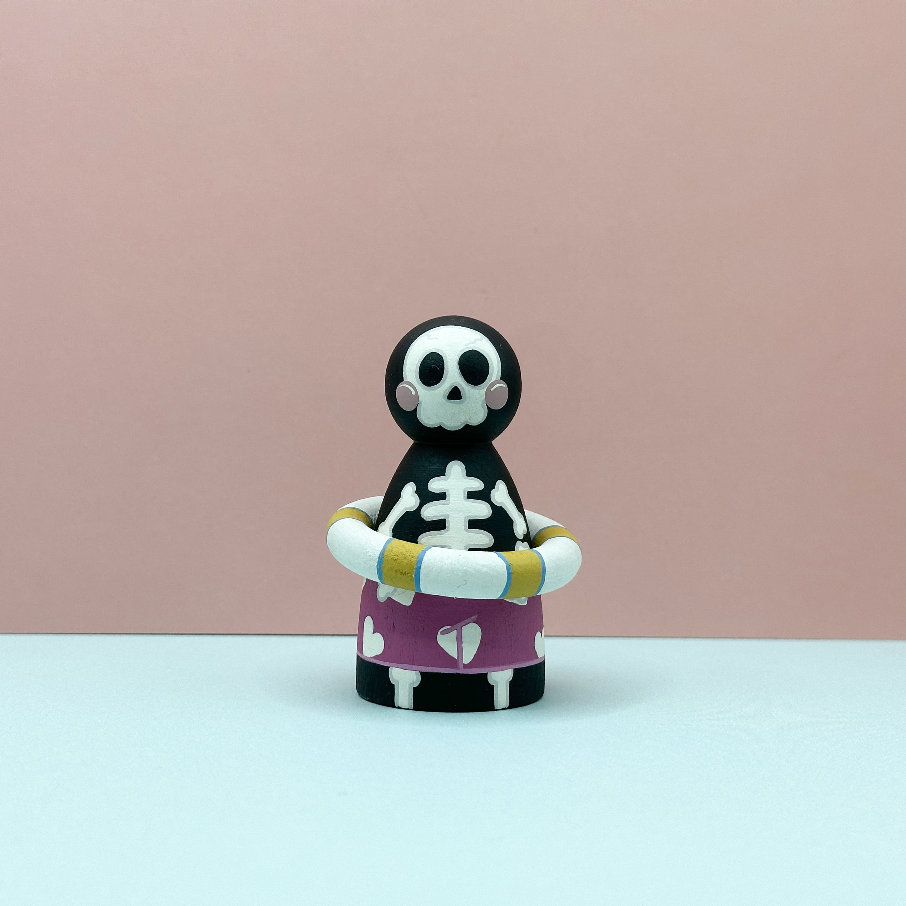 Skeleton figurine with a purple shirt and yellow arm bands on a pink background