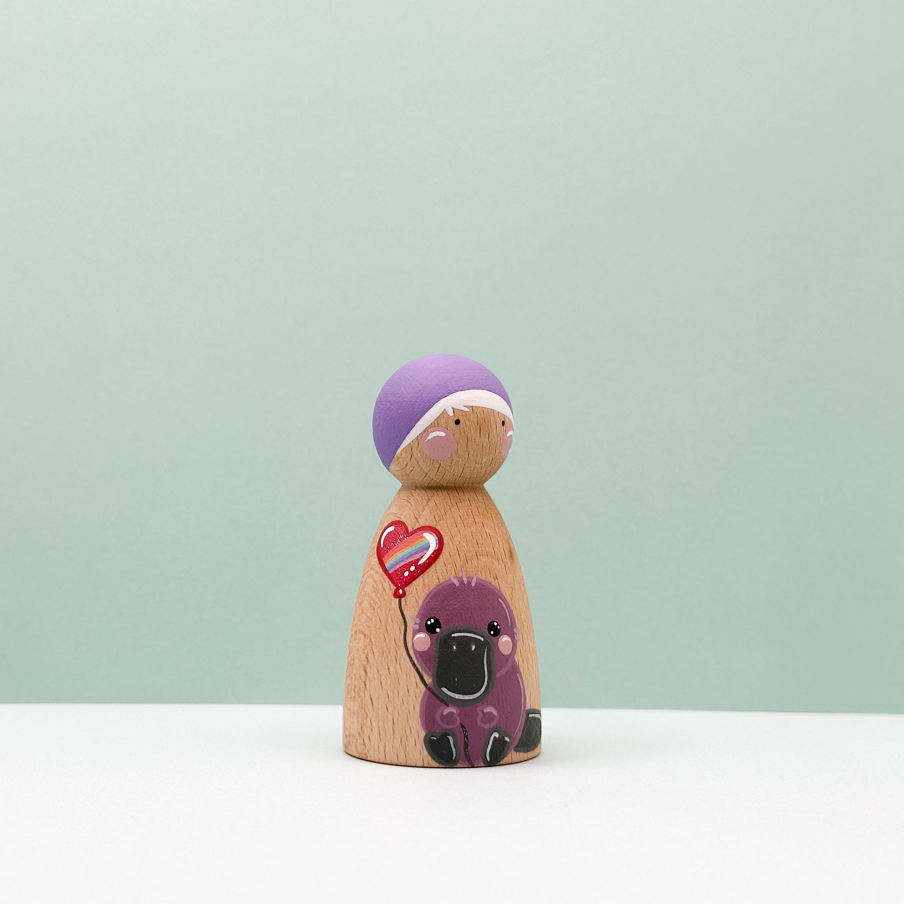 Wooden peg doll with a purple hat and heart-shaped balloon on a light green background