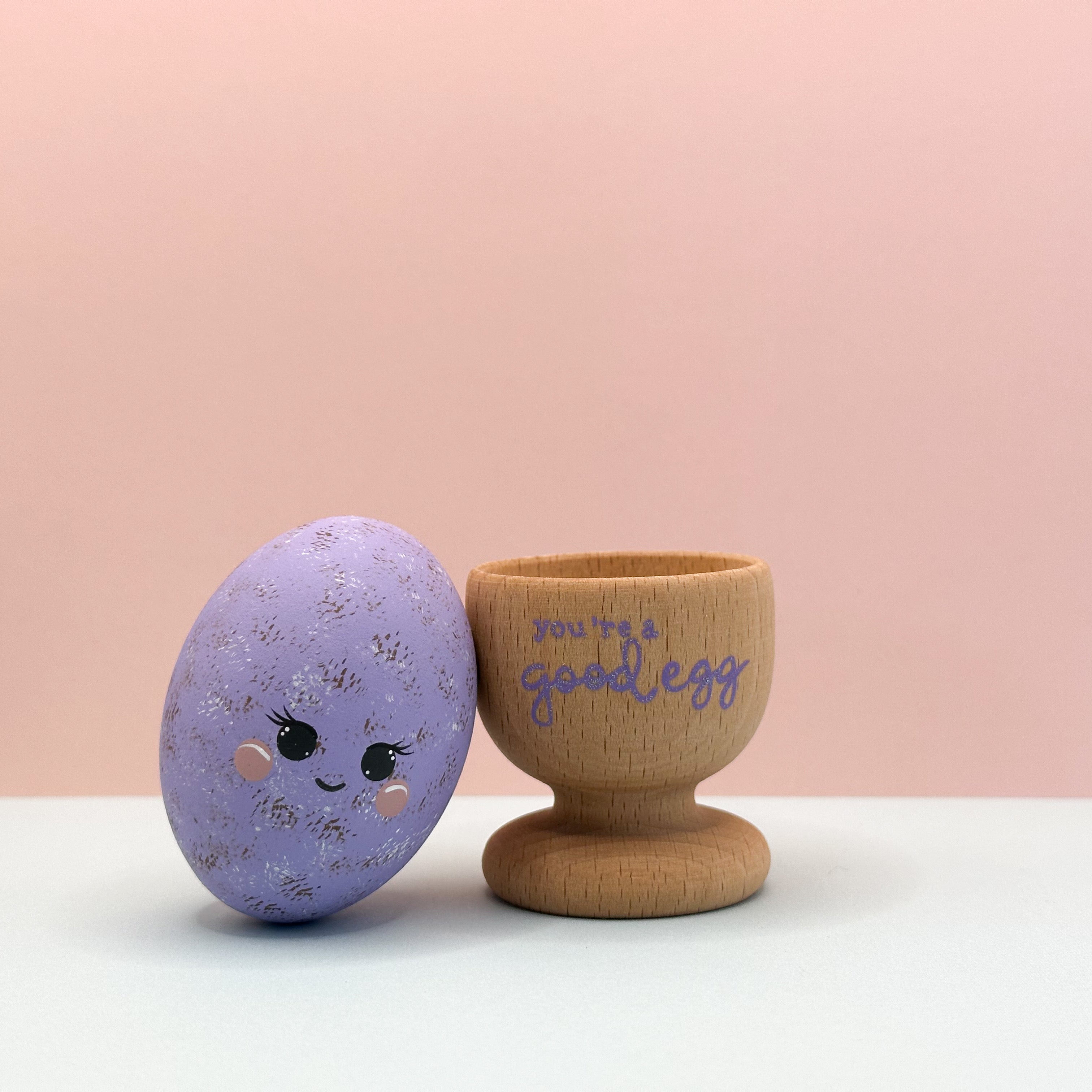 Decorative purple egg with a face and wooden egg cup with 'you're a good egg' text on a pink background