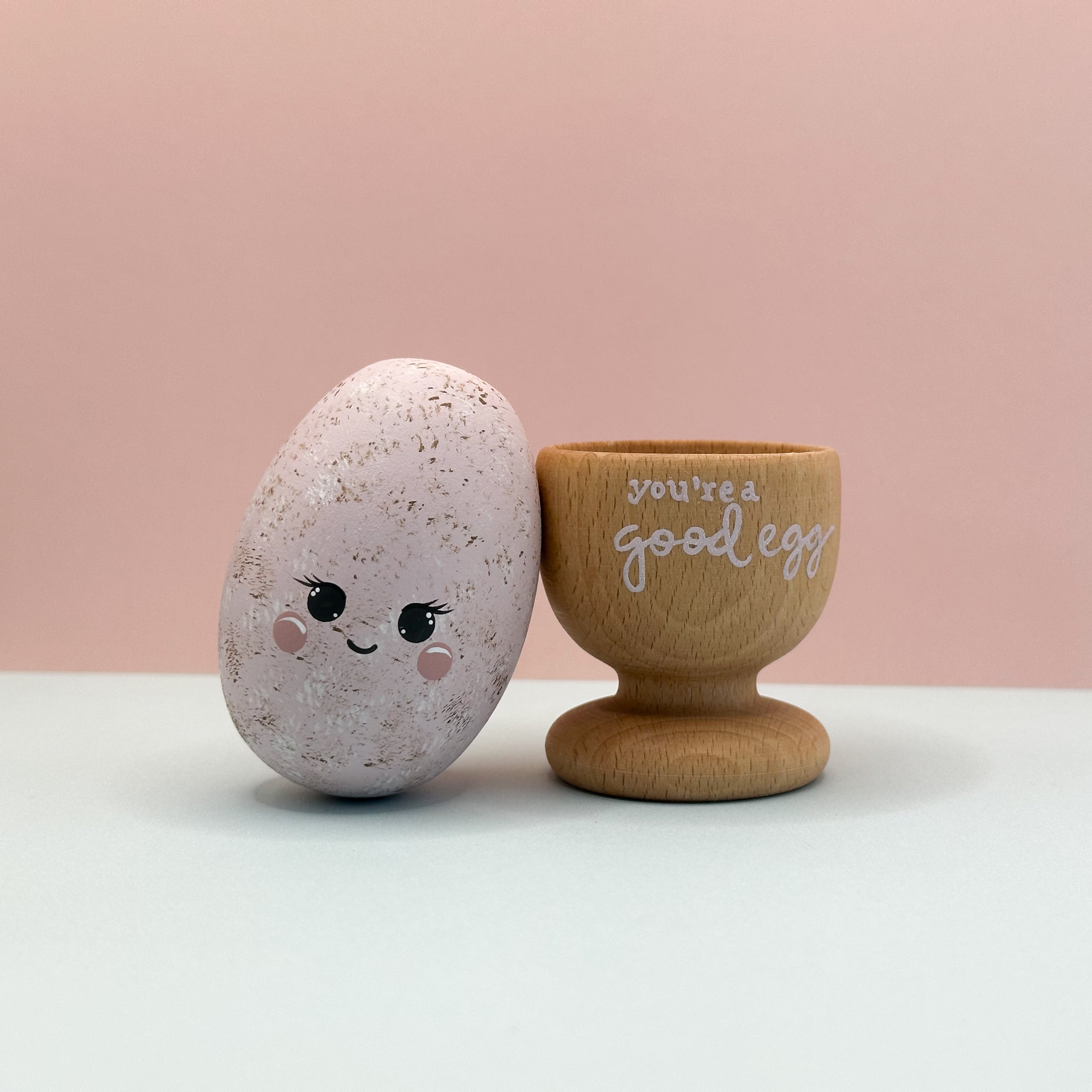 Decorative egg with a face and wooden cup with 'you're a good egg' text on a pink background