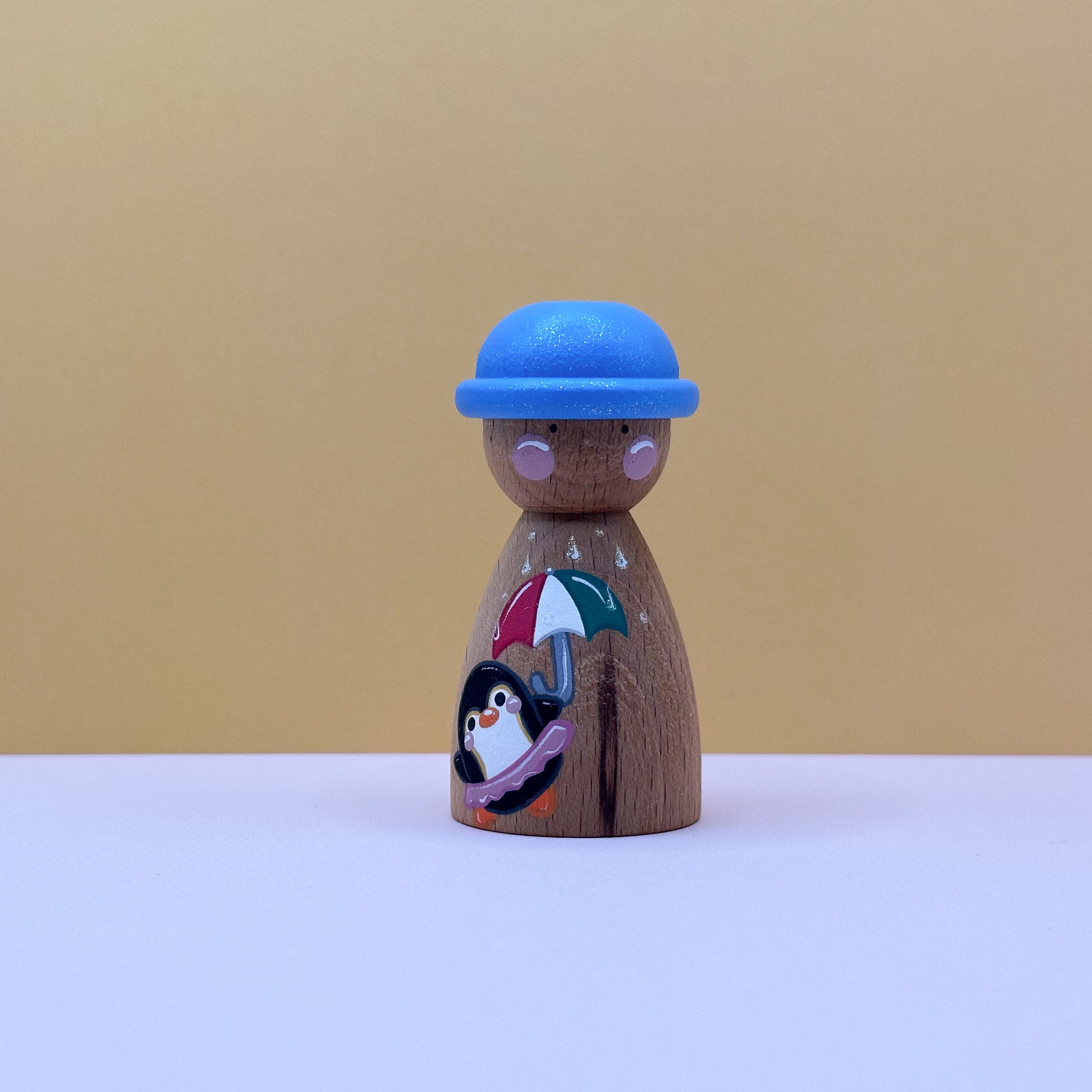 Wooden peg doll with a blue hat and umbrella design on a beige background