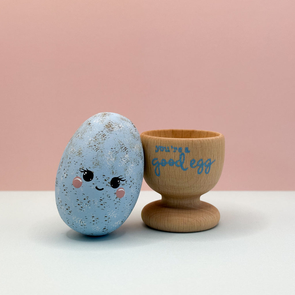 Blue speckled egg with a face and wooden stand with 'you're a good egg' text on a pink background