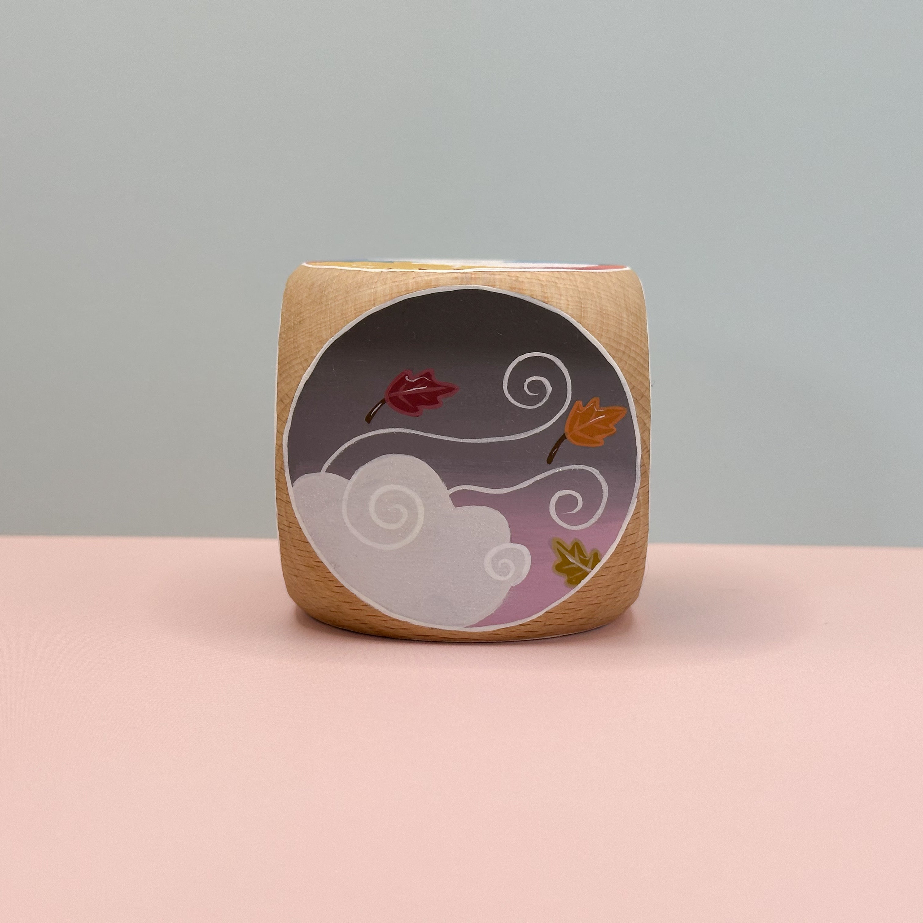 Decorative wooden cube featuring leaves and clouds on a pink surface.