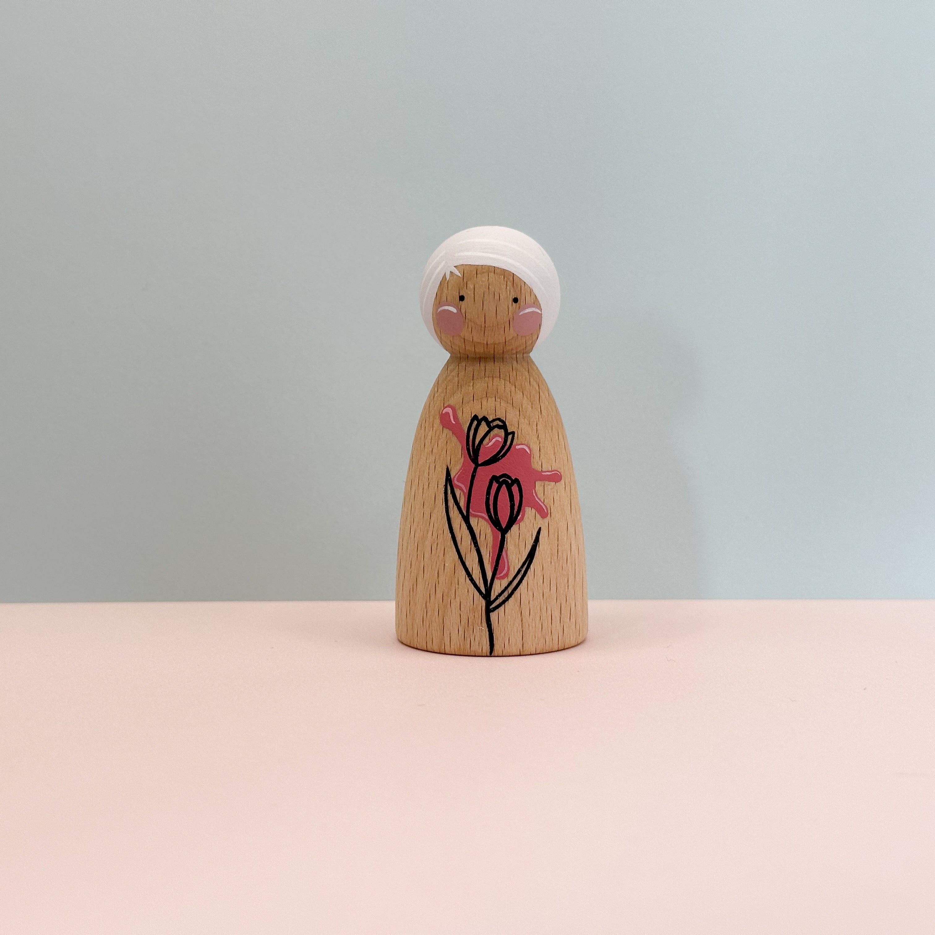 Wooden peg doll with a floral design on a light pink surface and light blue background