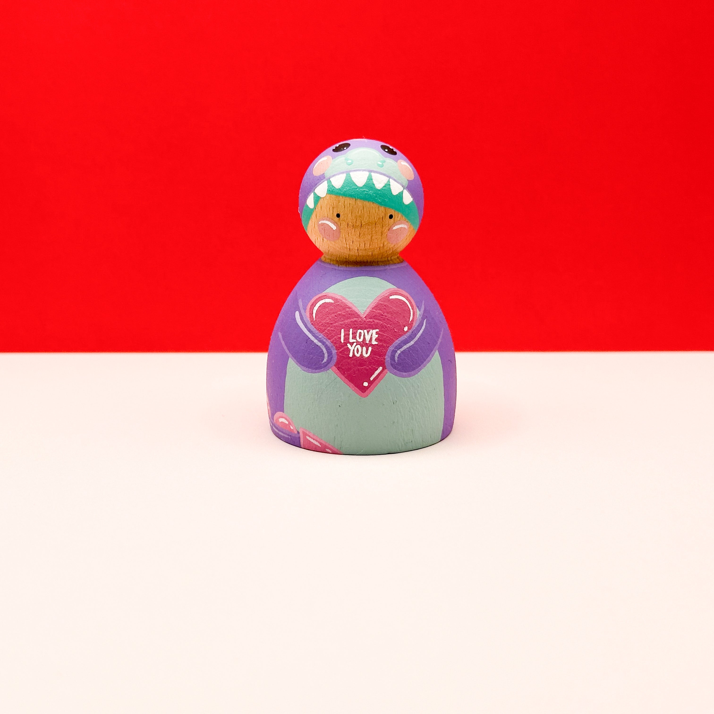 a chubby wooden peg doll dressed as a purple dinosaur holding a love heart on a red background