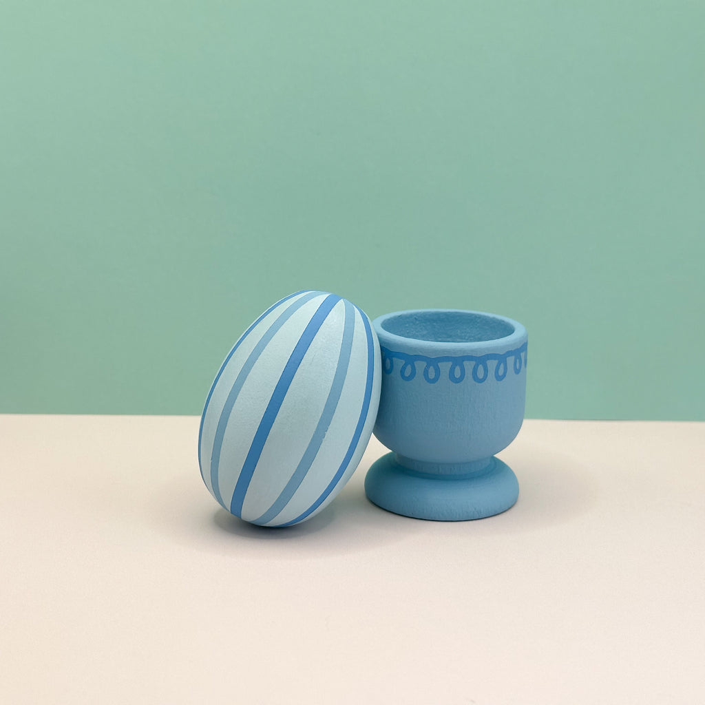Blue  egg with striped pattern and matching egg cup on a beige surface with a green background