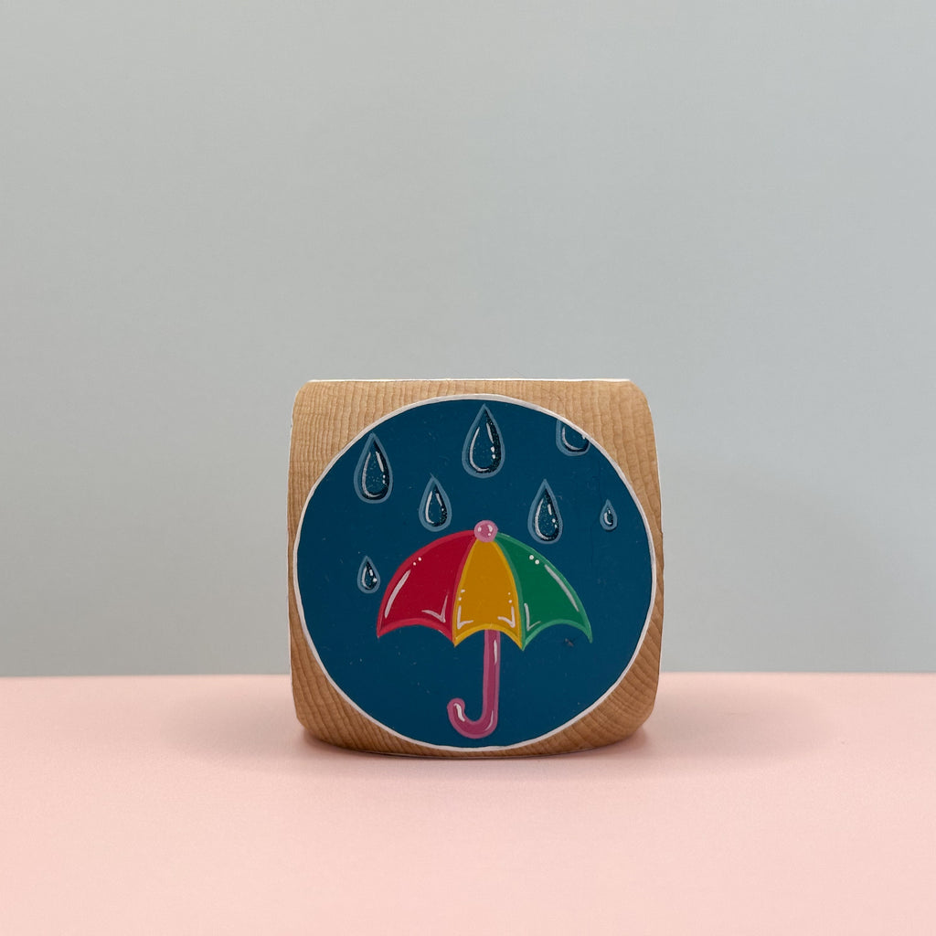 Wooden block with a colorful umbrella design on a blue background, placed on a pink surface with a gray background.