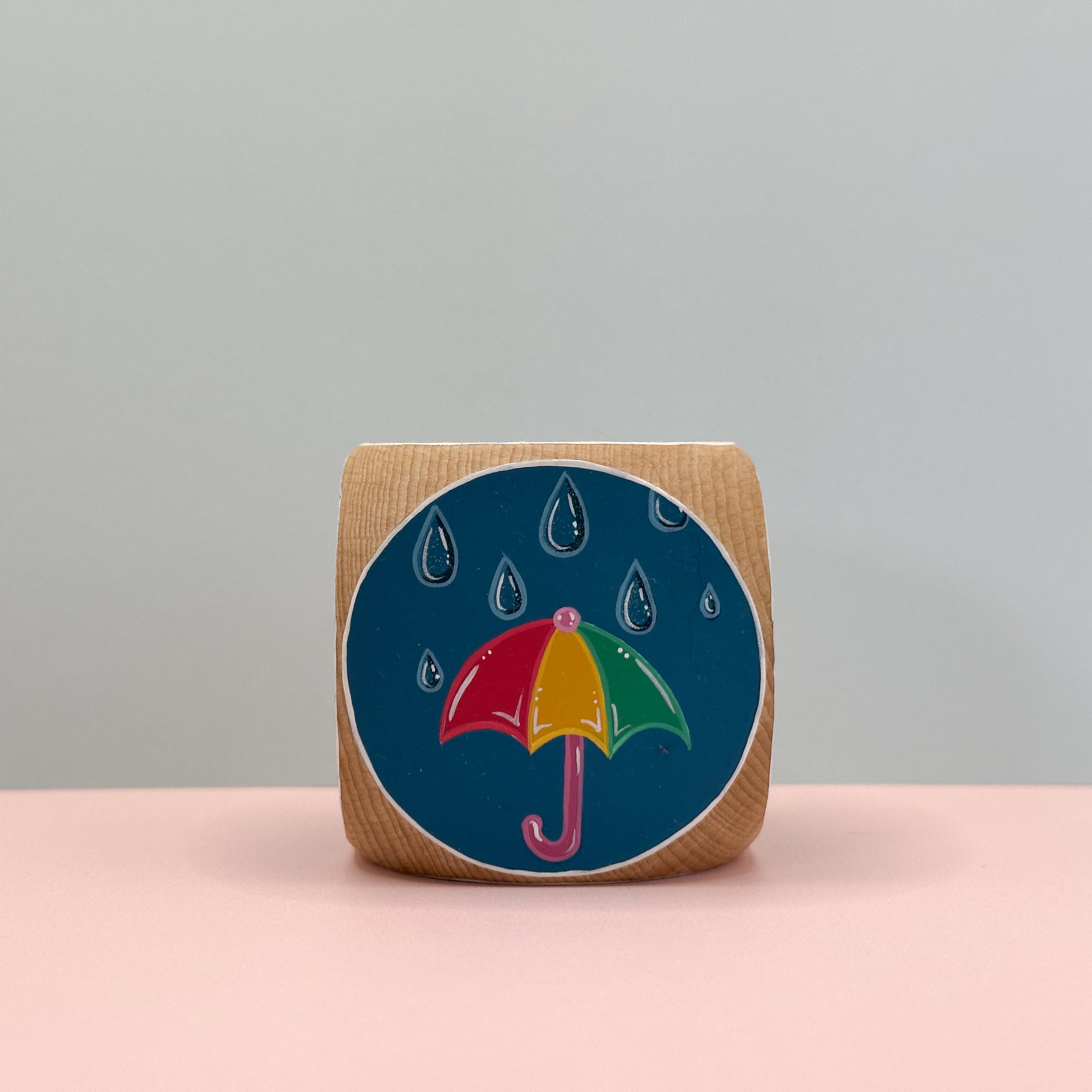Wooden block with a colorful umbrella design on a blue background, placed on a pink surface with a gray background.
