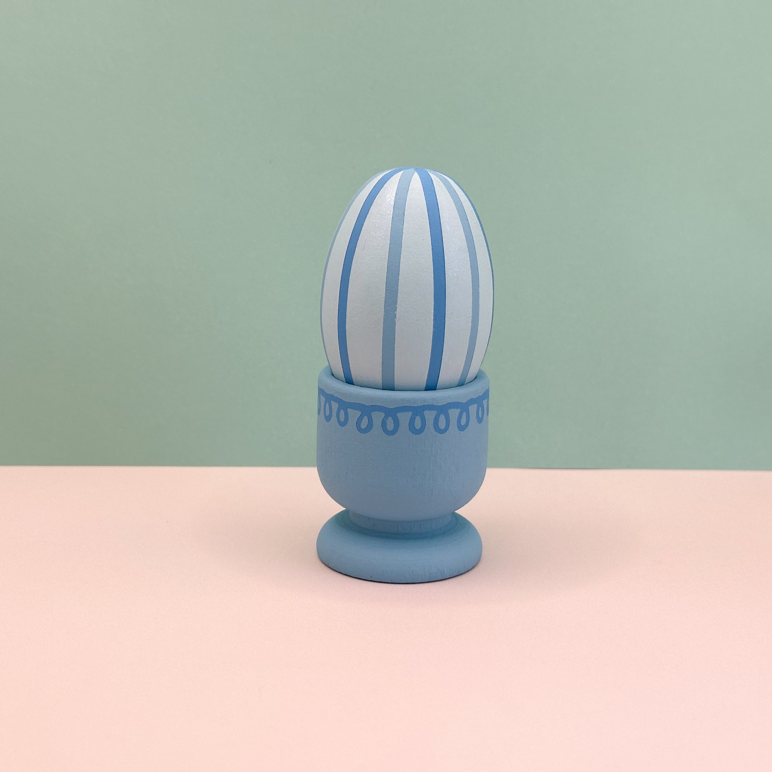 Decorative egg with blue and white stripes in a matching blue holder on a pink surface with a green background