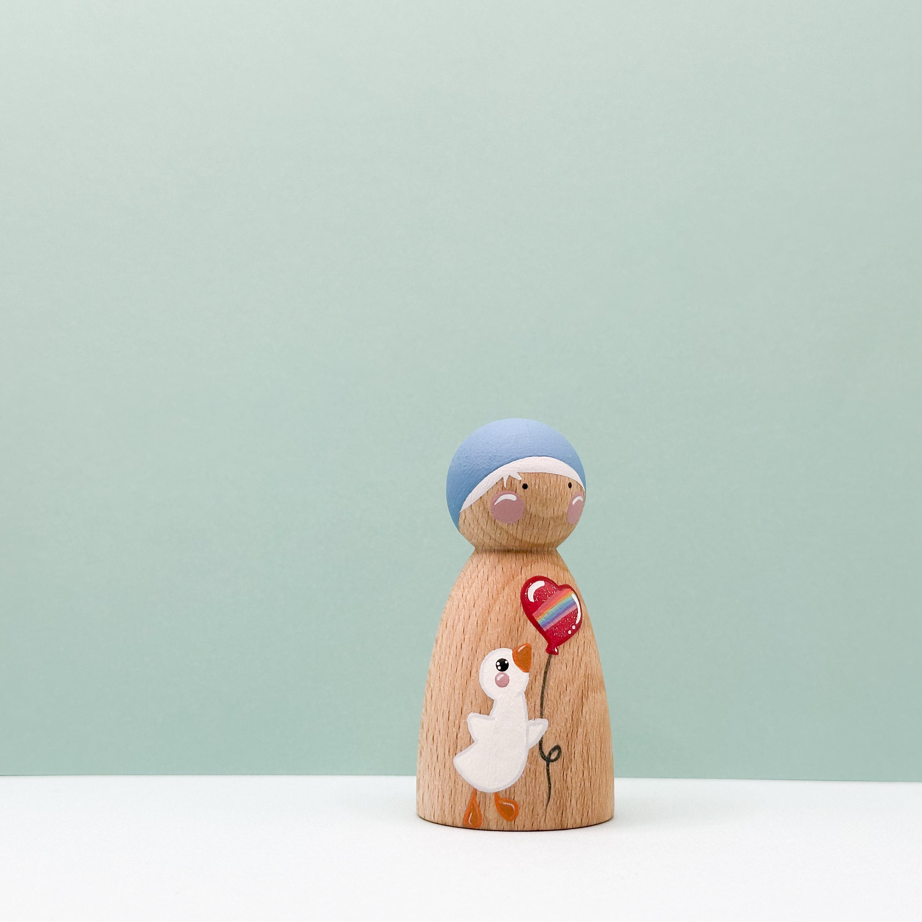 Wooden peg doll with a blue hat and red heart balloon on a light green background