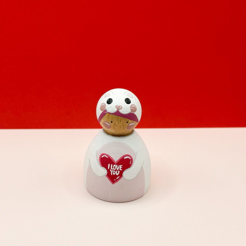 a chubby wooden peg doll dressed as a cat holding a love heart on a red background