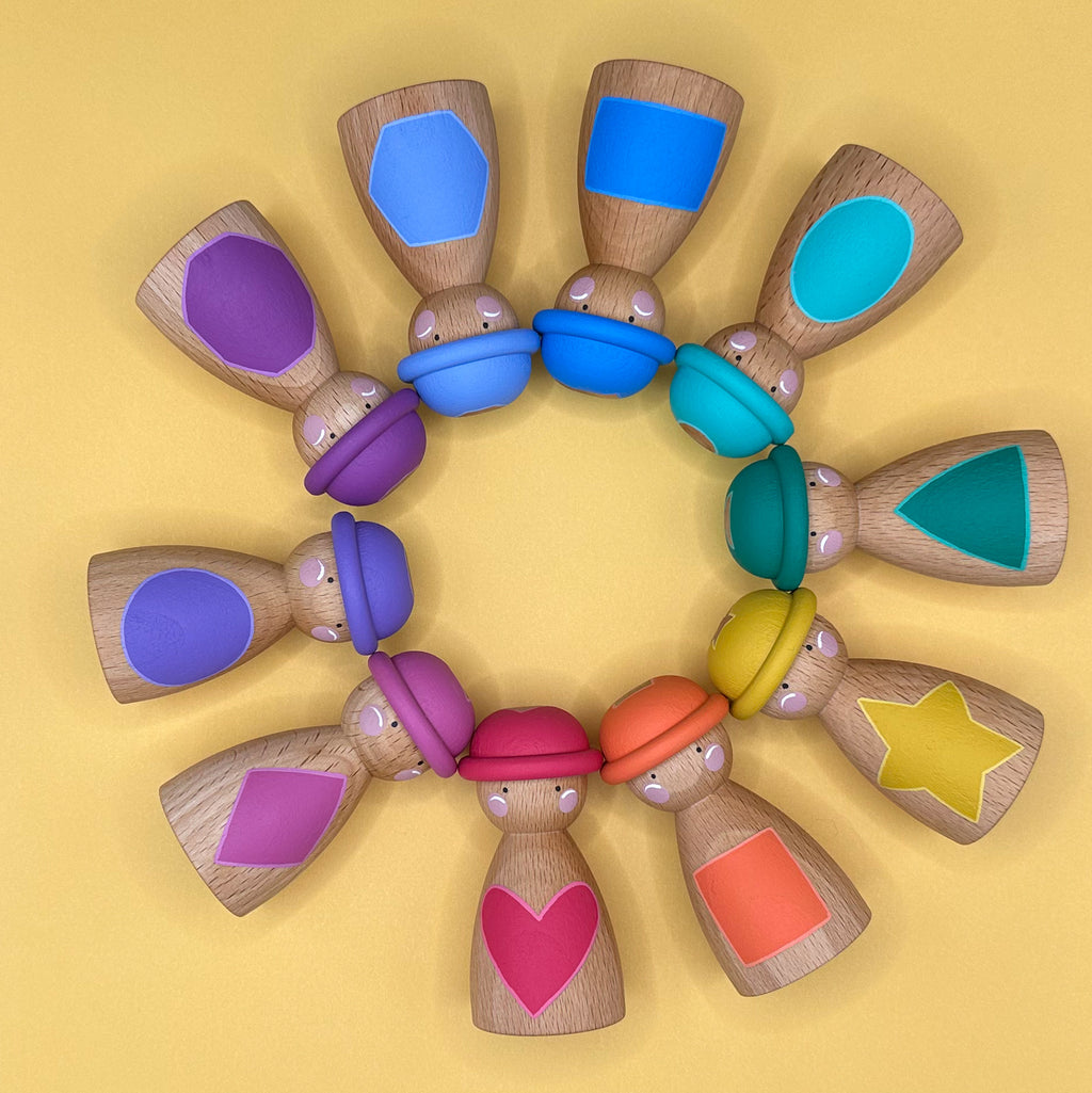 Wooden Dolls with colorful hats arranged in a circle on a yellow background. The dolls have colourful painted shapes on their tummies. 